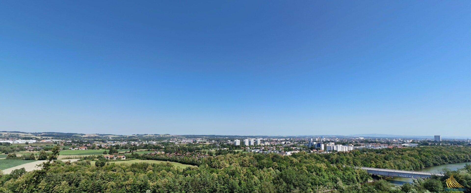 Weitläufiger Panoramablick über eine grüne Landschaft mit Stadtansicht und Fluss im Hintergrund.