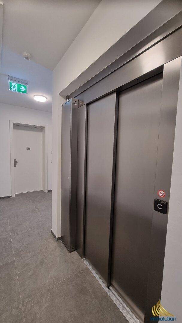 Moderner Aufzug in einem hellen Flur mit Fliesenboden und Notausgangsschild.