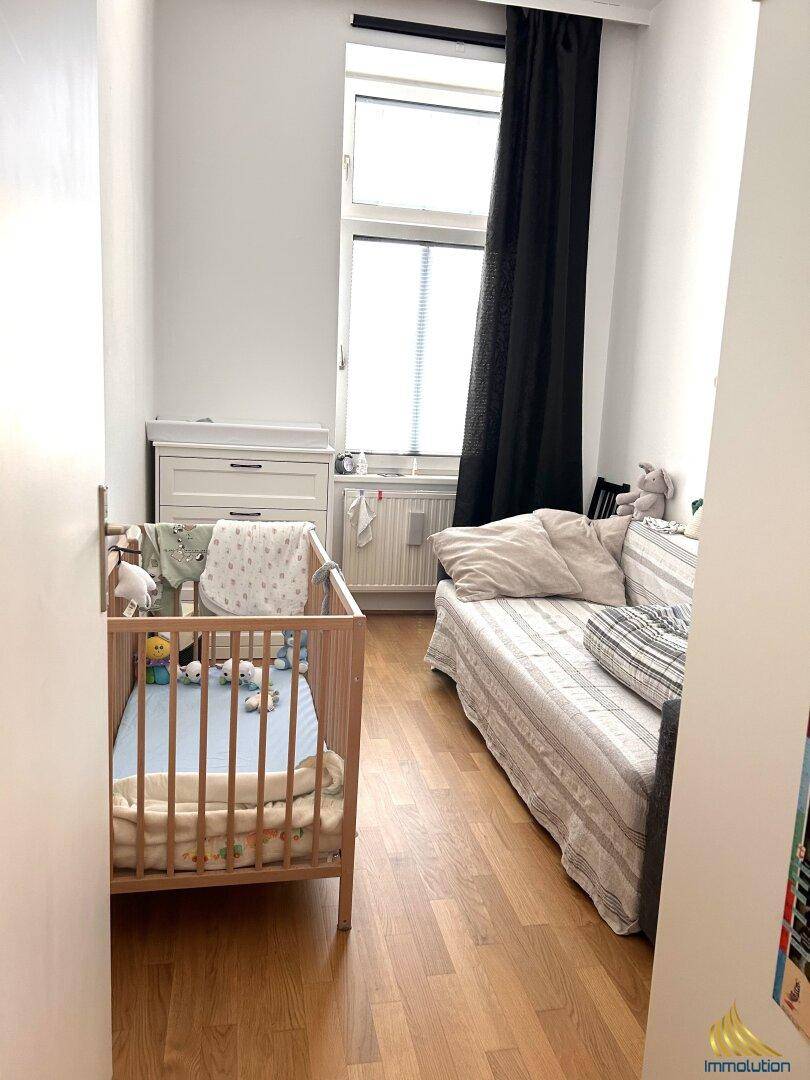 Helles Kinderzimmer mit Holzboden, Babybett und einer Liege am Fenster.