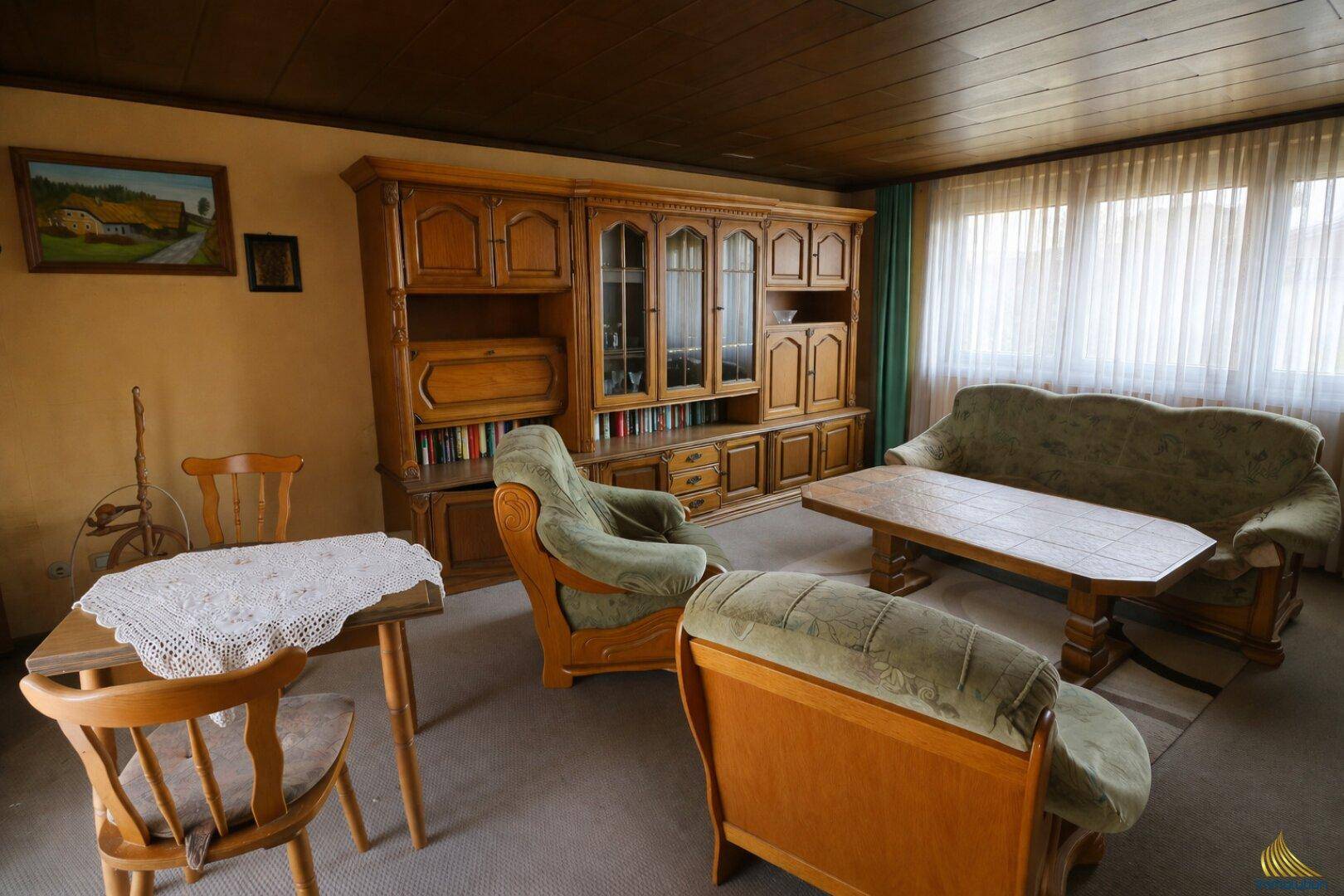 Traditionelles Wohnzimmer mit Holzdecke, Teppichboden und rustikalen Holzmöbeln am Fenster.