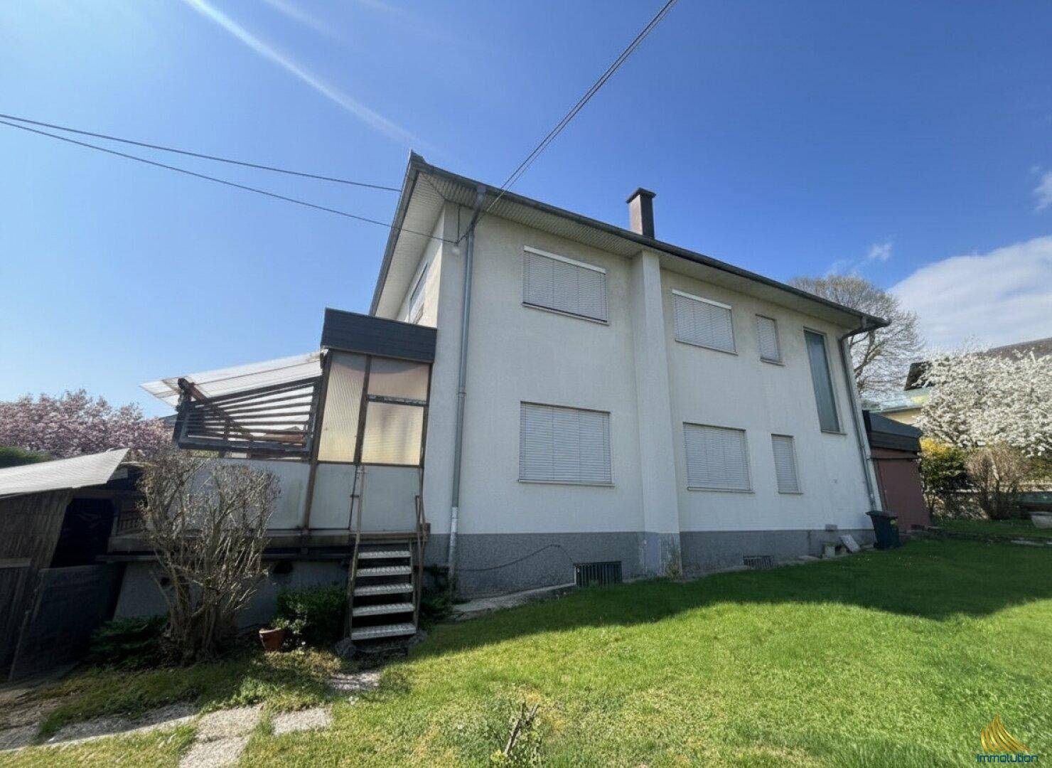 Rückseite des Hauses mit grünem Garten, Rasenfläche und einem verglasten Anbau.