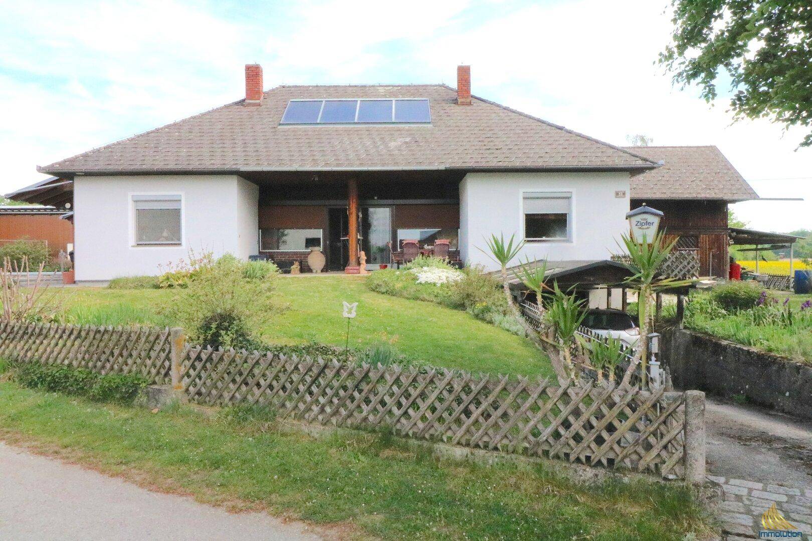 Vorderansicht des Hauses mit überdachter Terrasse, Garten und Solarpaneelen auf dem Dach.