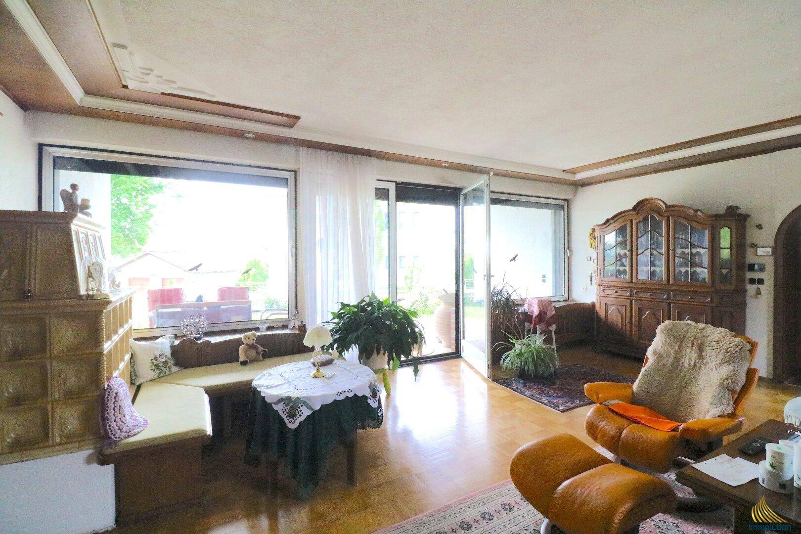 Lichtdurchflutetes Wohnzimmer mit Zugang zur Terrasse, Kachelofen und gemütlicher Sitzecke.