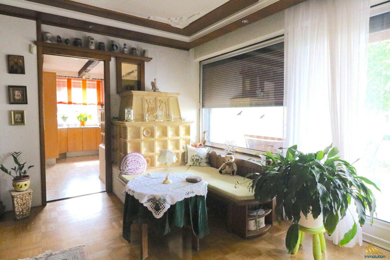 Helles Wohnzimmer mit Kachelofen, Parkettboden und Zugang zur Küche.