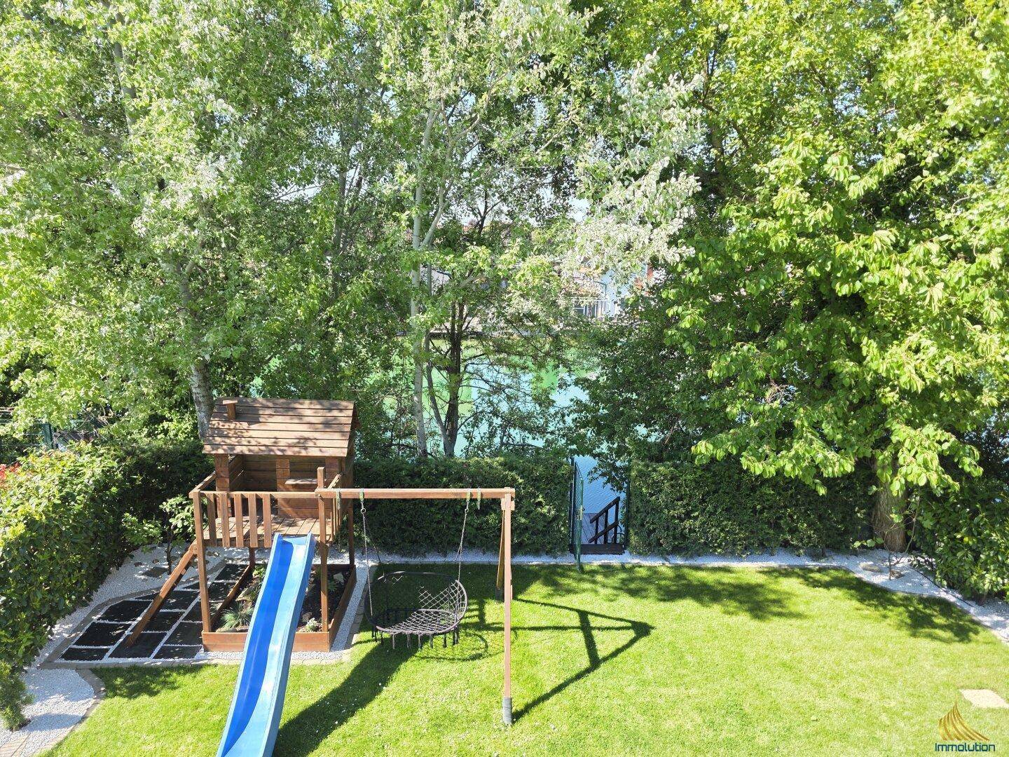 Blick von oben auf den Garten mit Spielplatz, Rutsche und Wasserblick im Hintergrund.