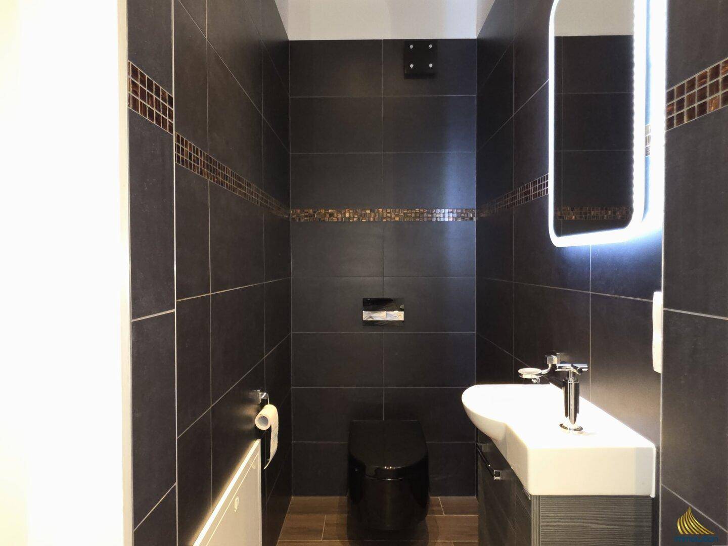Modernes Gäste-WC mit dunklen Fliesen, Wand-WC und beleuchtetem Spiegel.