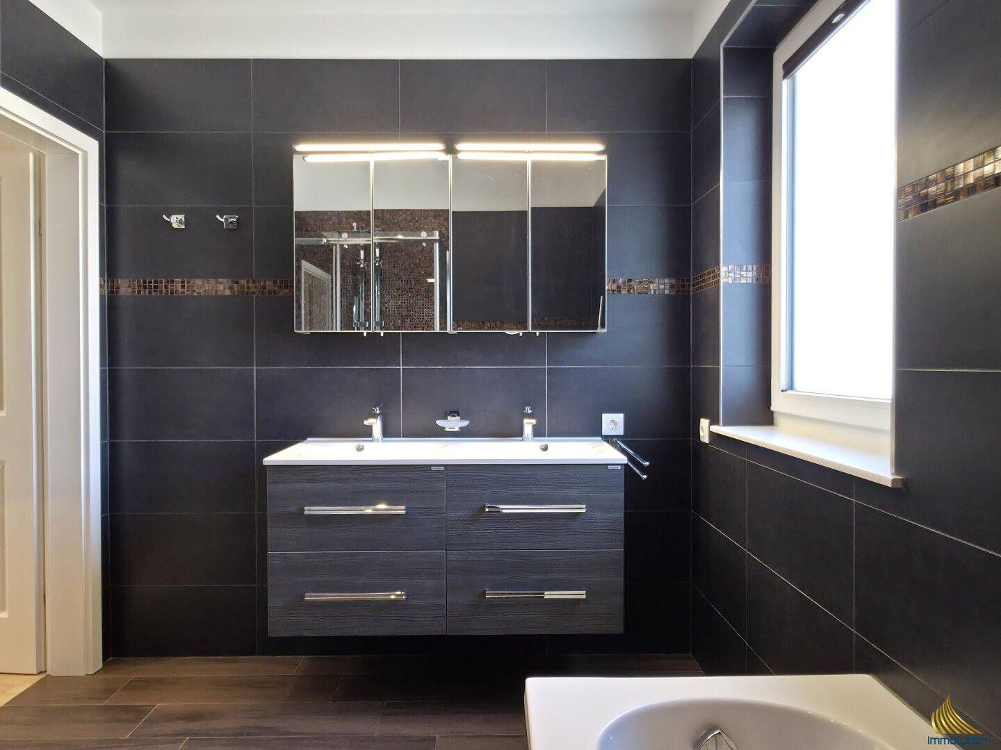 Modernes Badezimmer mit Doppelwaschbecken, dunklen Fliesen und Fenster.