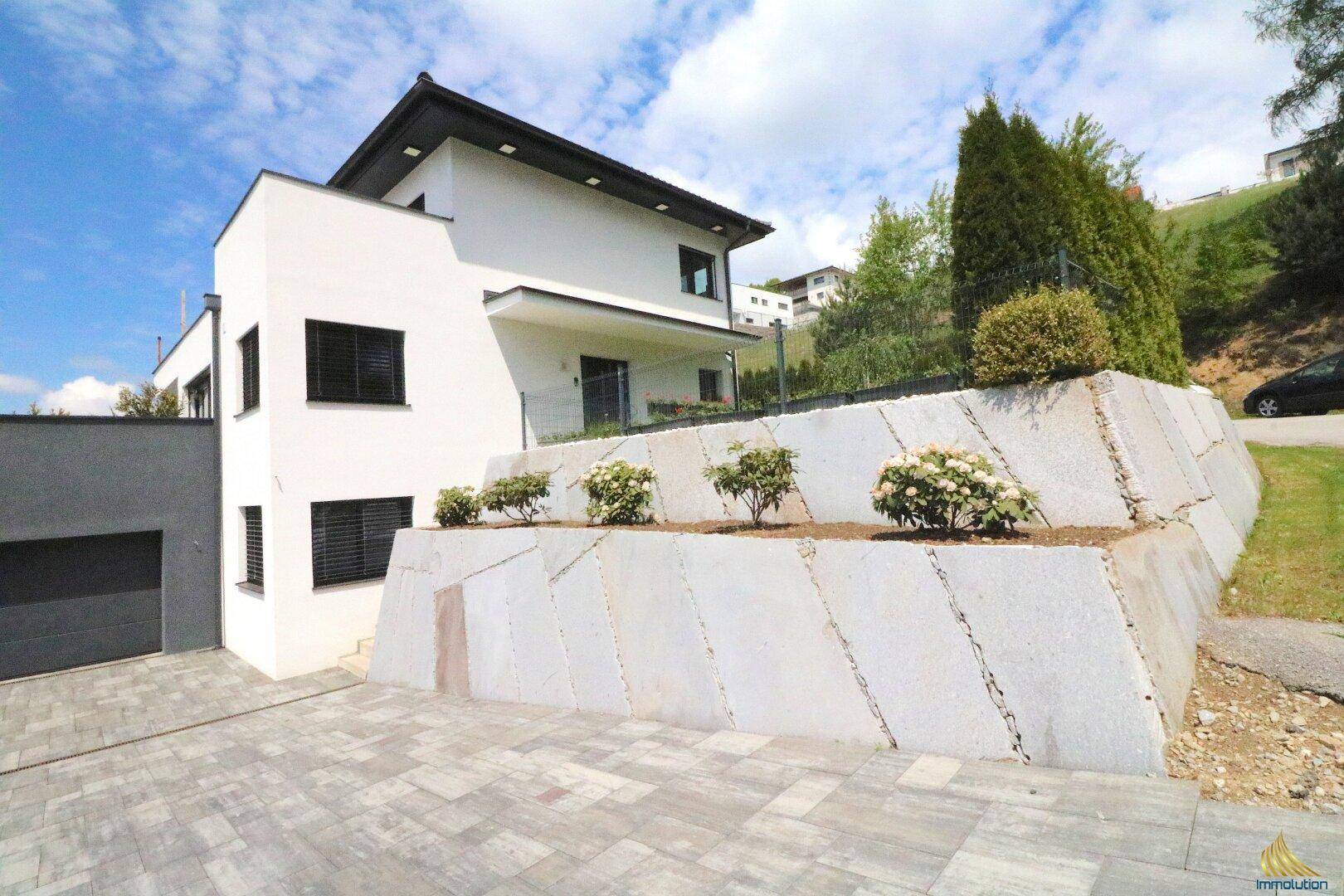 Seitenansicht der modernen Villa mit gepflasterter Auffahrt und einer begrünten Stützmauer.