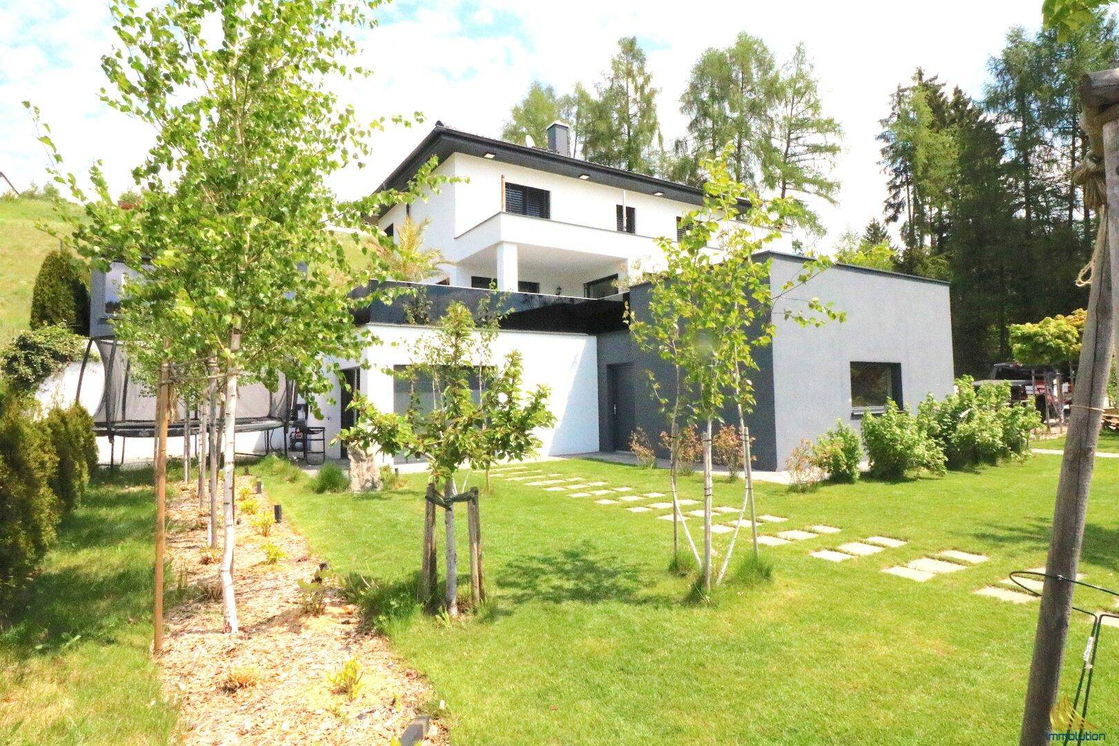 Blick vom Garten auf die moderne Villa mit weißer Fassade, grüner Rasenfläche und jungen Bäumen.