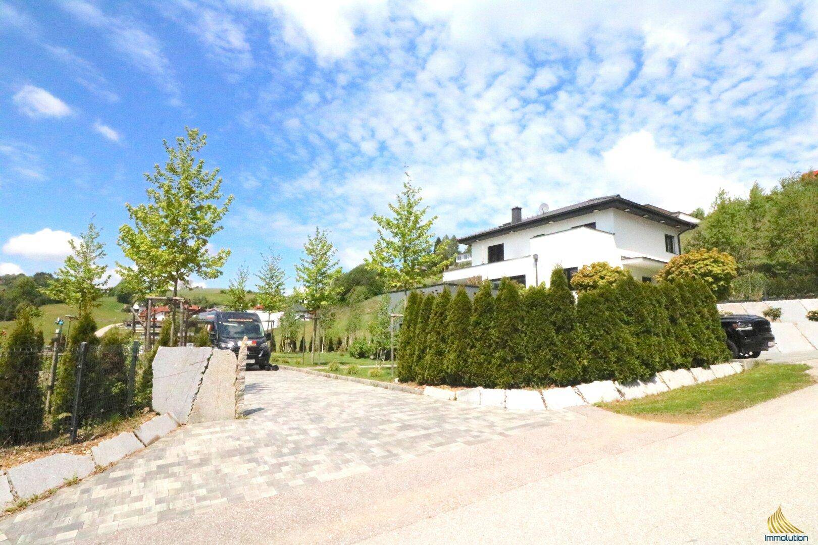 Zufahrt zur modernen Villa von der Straße aus, mit gepflasterter Auffahrt und hohen Hecken.