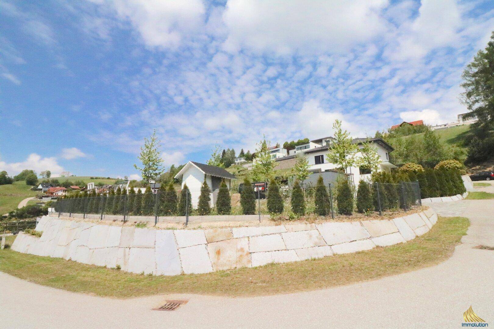 Weitläufiger Blick von der Straße auf die moderne Villa, umgeben von grünen Hügeln und einer Stützmauer.