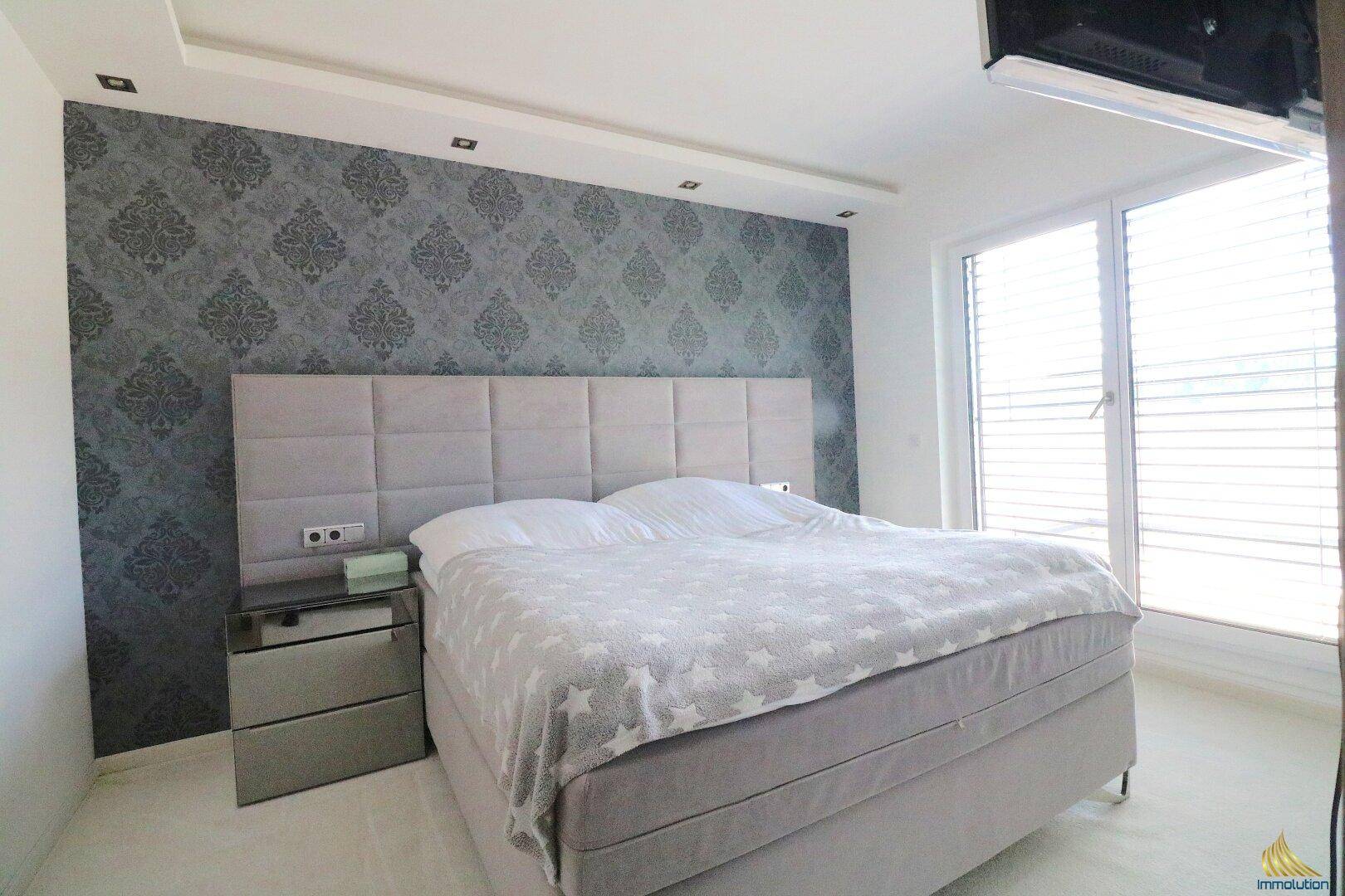 Gemütliches Schlafzimmer mit großem Boxspringbett, gemusterter Tapete und viel Tageslicht.