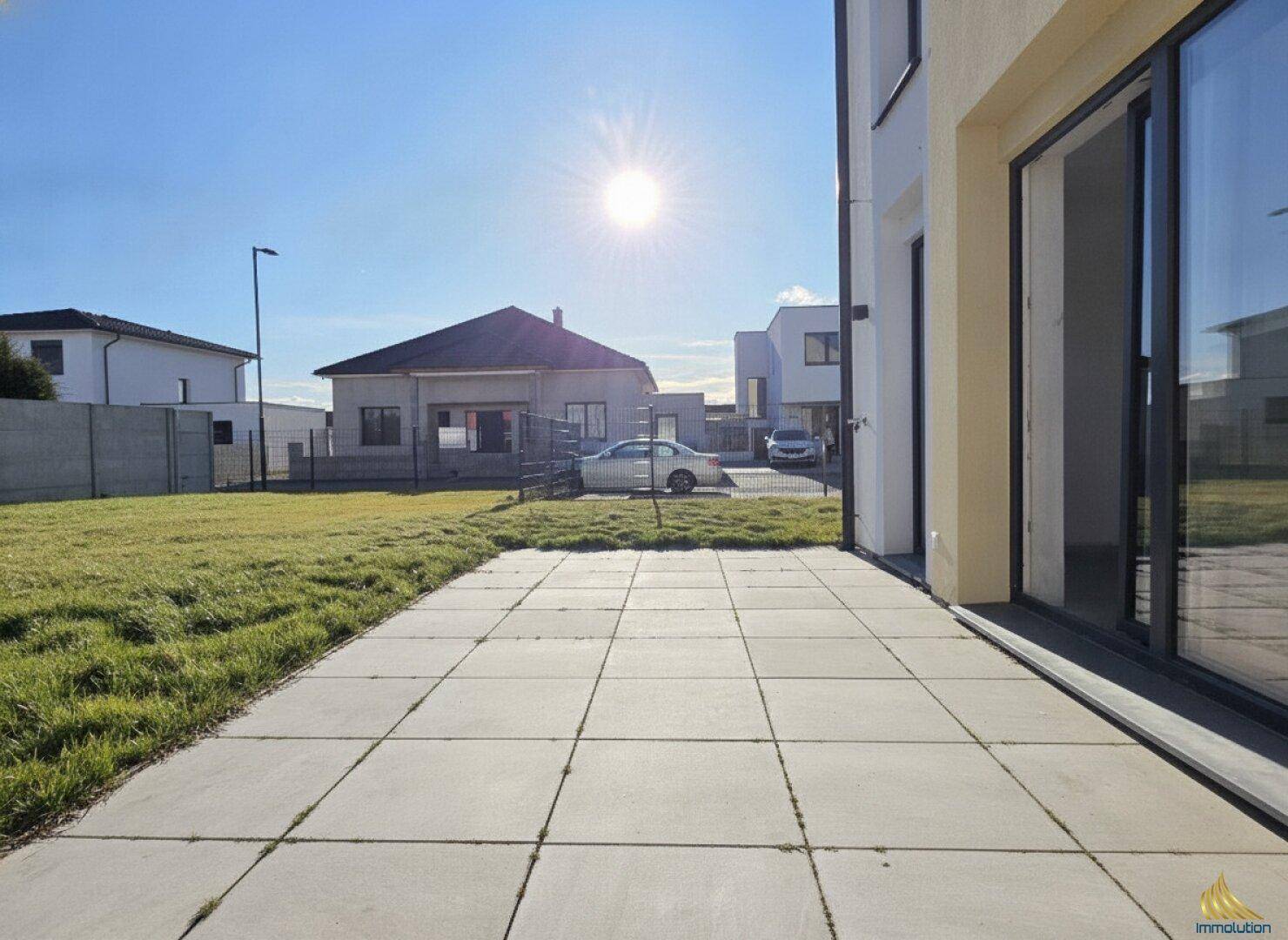 Sonnige Terrasse mit großformatigen Fliesen und angrenzendem Rasenbereich am Neubau.