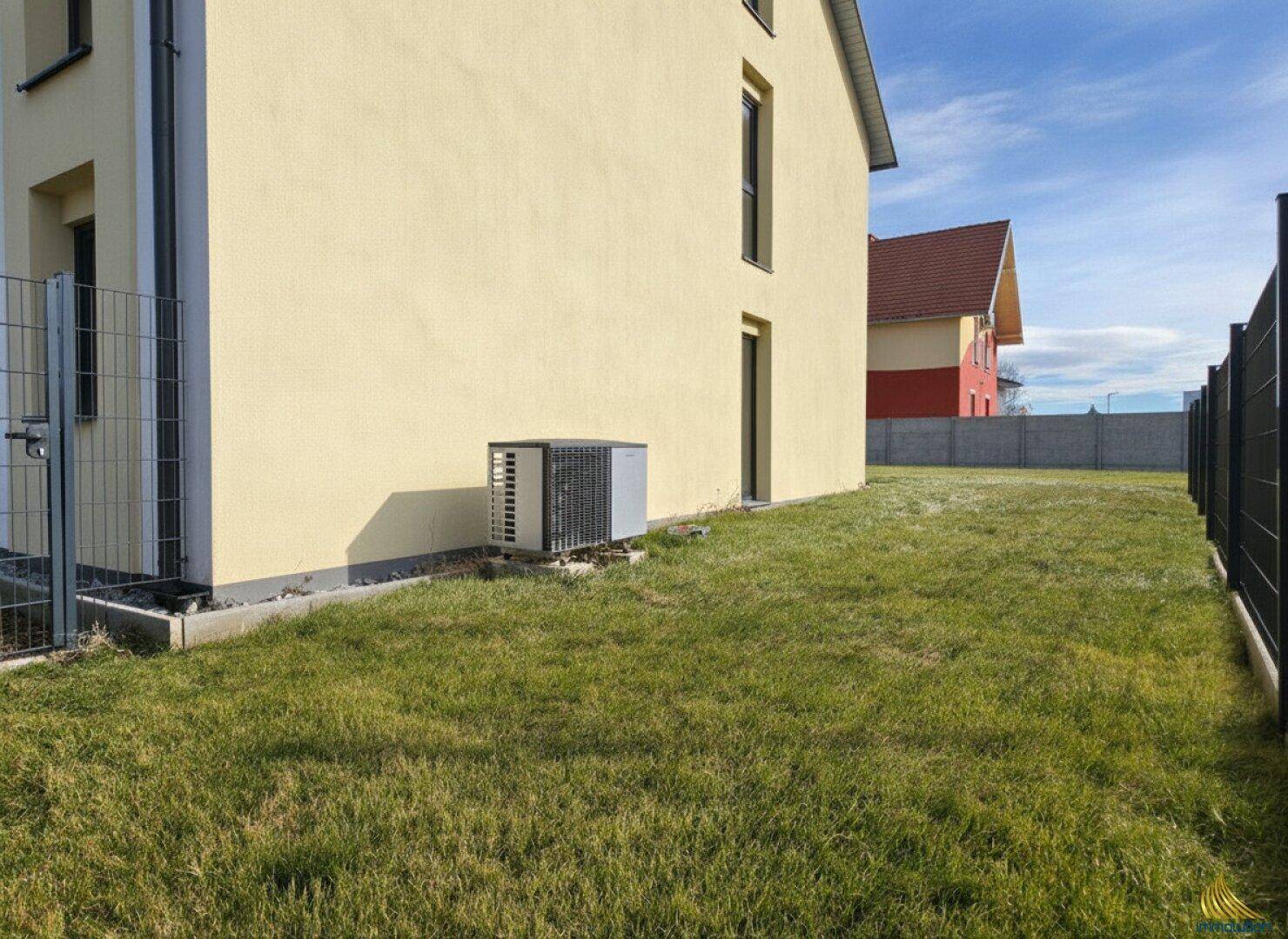 Seitenansicht des Hauses mit installierter Wärmepumpe und schmalem Rasenstreifen.