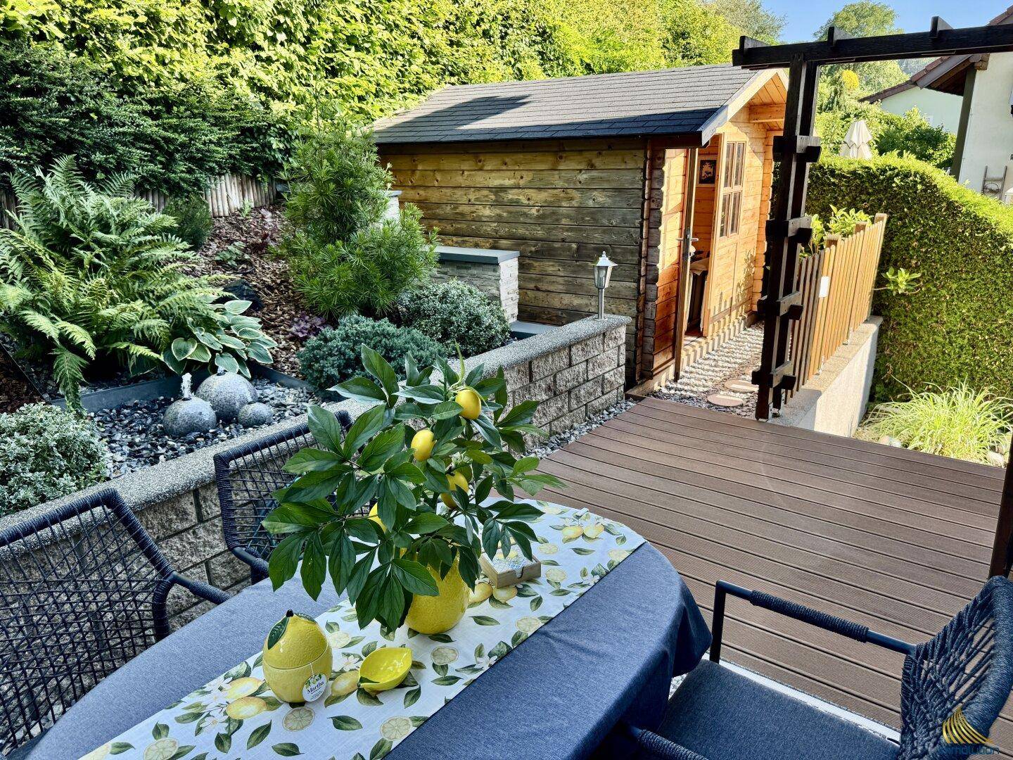 Gemütlicher Essbereich im Freien auf einer Holzterrasse mit Gartenhaus und viel Grün.