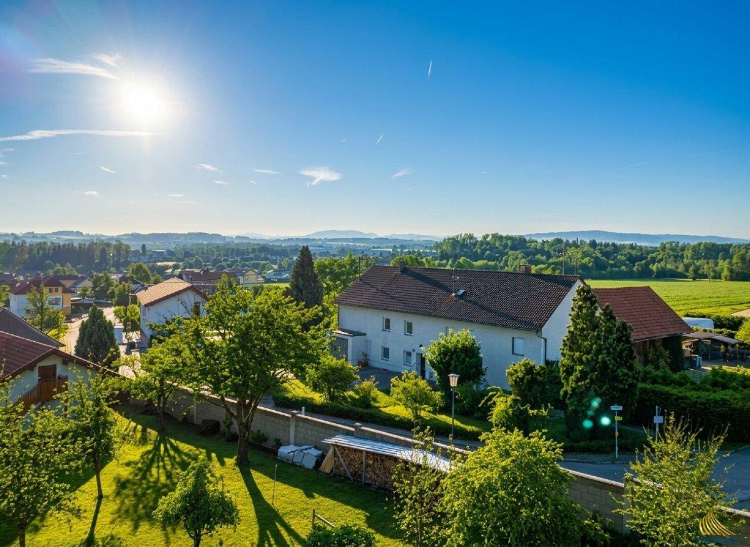 Weitläufige Ansicht einer Wohngegend mit viel Grün, Feldern und Bergen unter strahlend blauem Himmel.