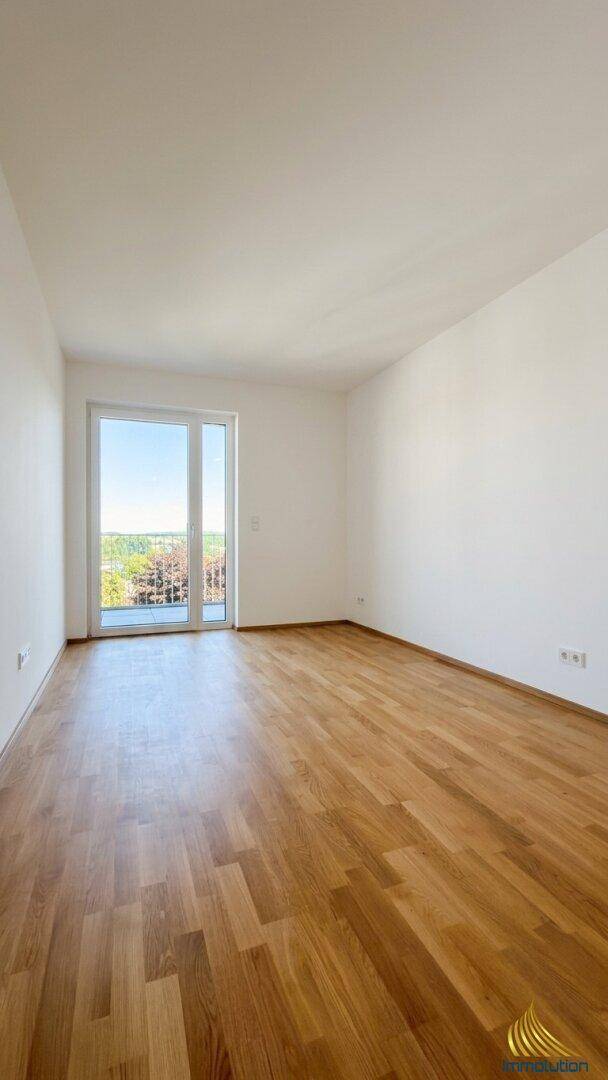 Heller, leerer Raum mit Holzboden und großem Fenster, das Zugang zu einem kleinen Balkon bietet.