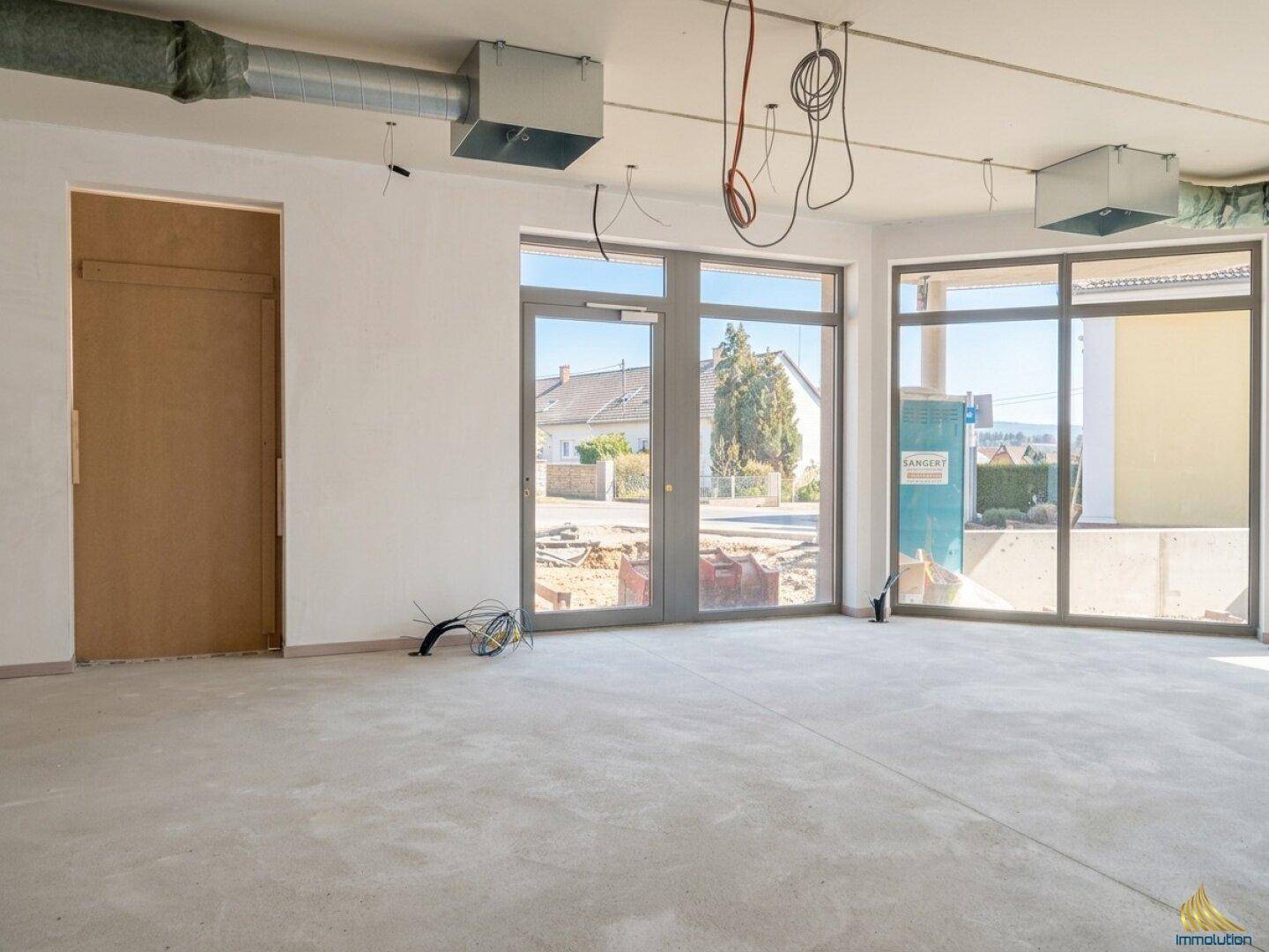 Unfertiger Gewerberaum mit großen Fenstern, Betonboden und sichtbaren Installationen.