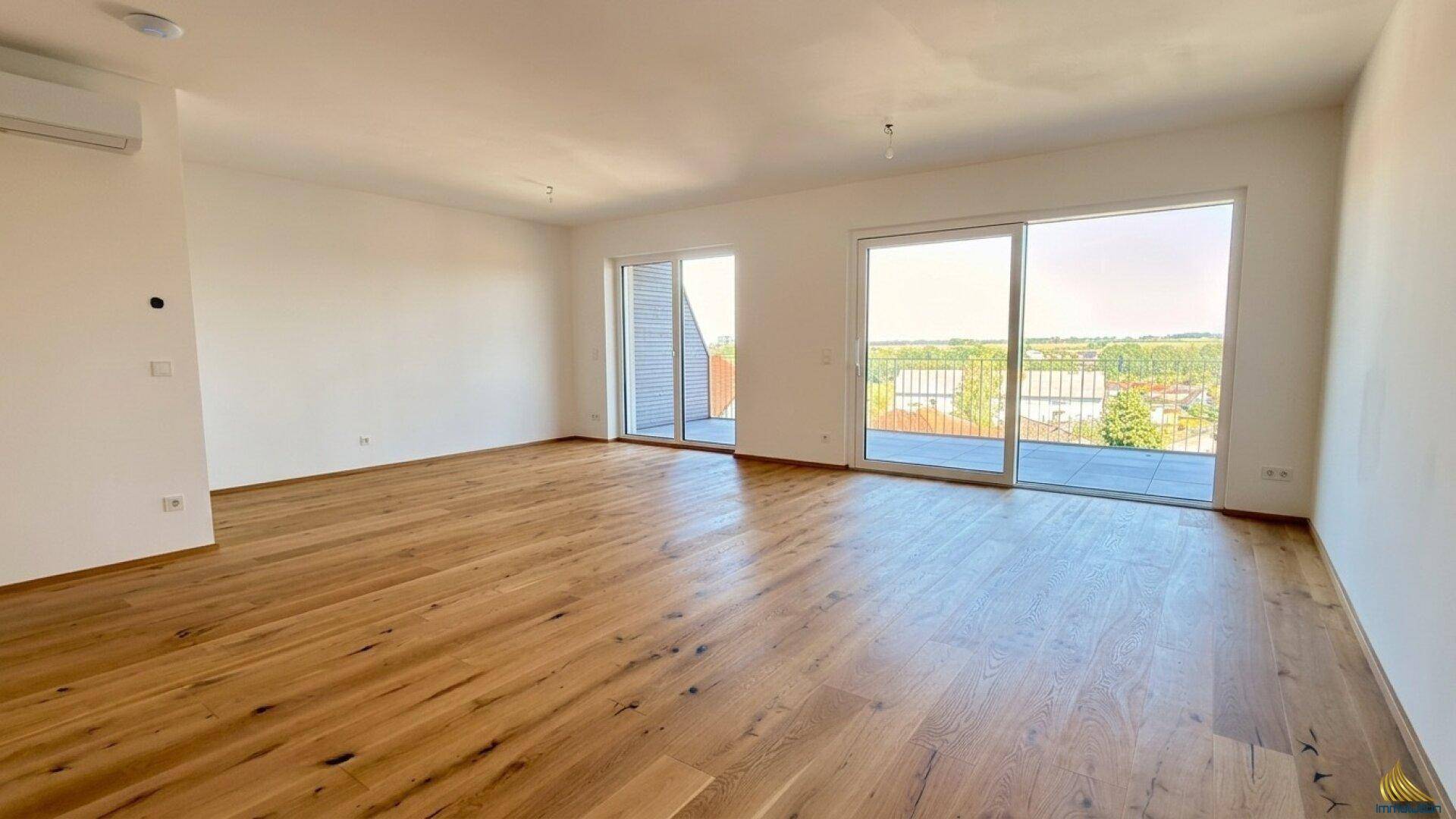 Helles, leeres Wohnzimmer mit breiten Holzdielen, großen Schiebetüren zum Balkon und Klimaanlage.