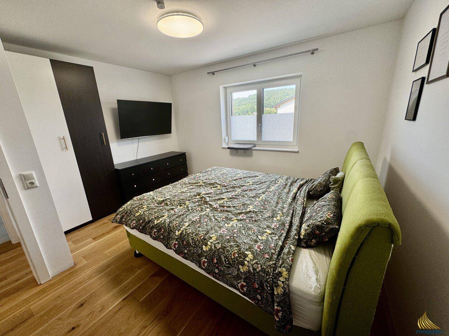 Schlafzimmer mit grünem Bett, dunklem Kleiderschrank und Holzboden, hell und freundlich.
