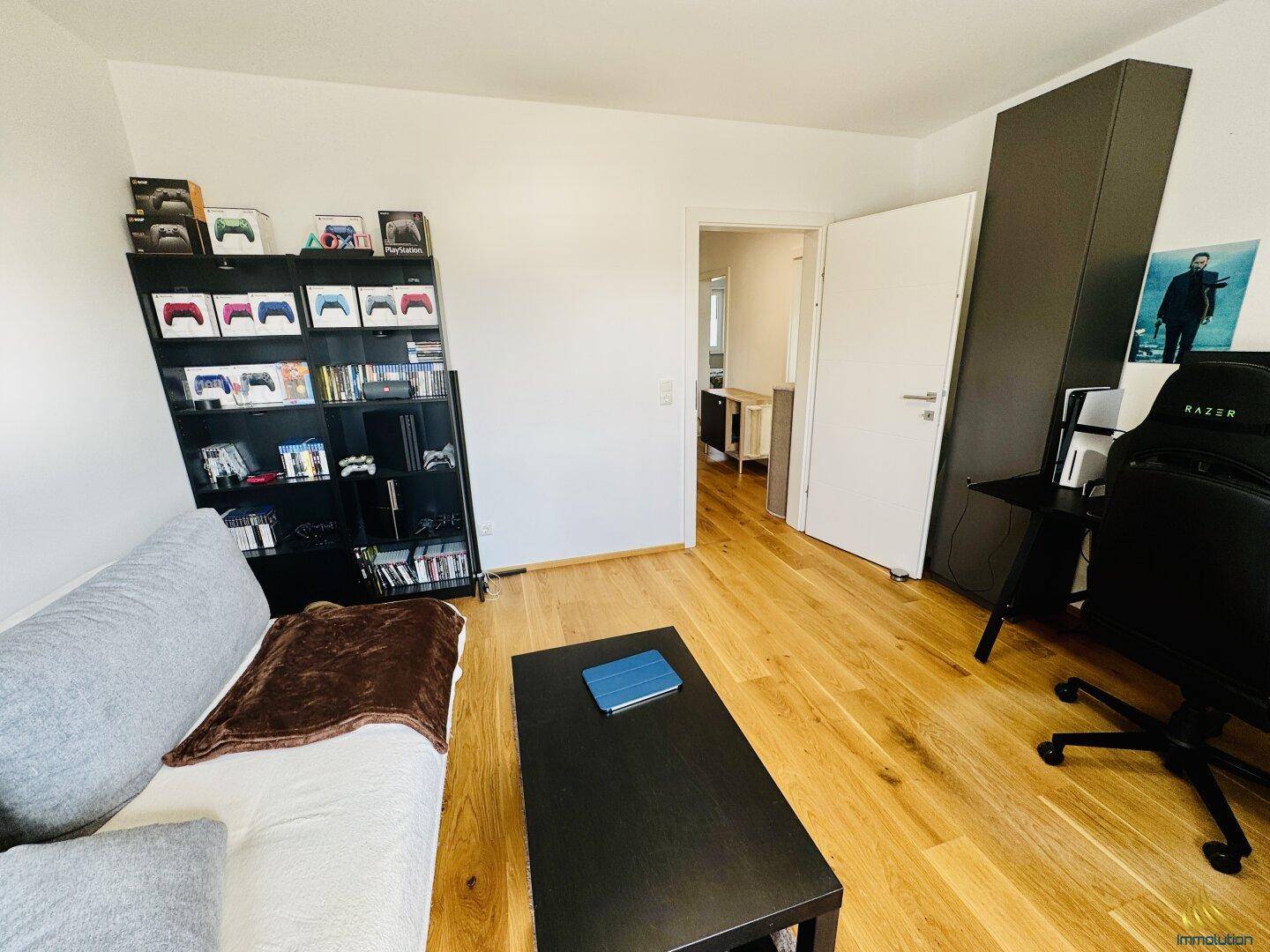 Helles Zimmer mit Holzboden, Sofa, Bücherregal und Gaming-Stuhl, ideal als Büro oder Hobbyraum.