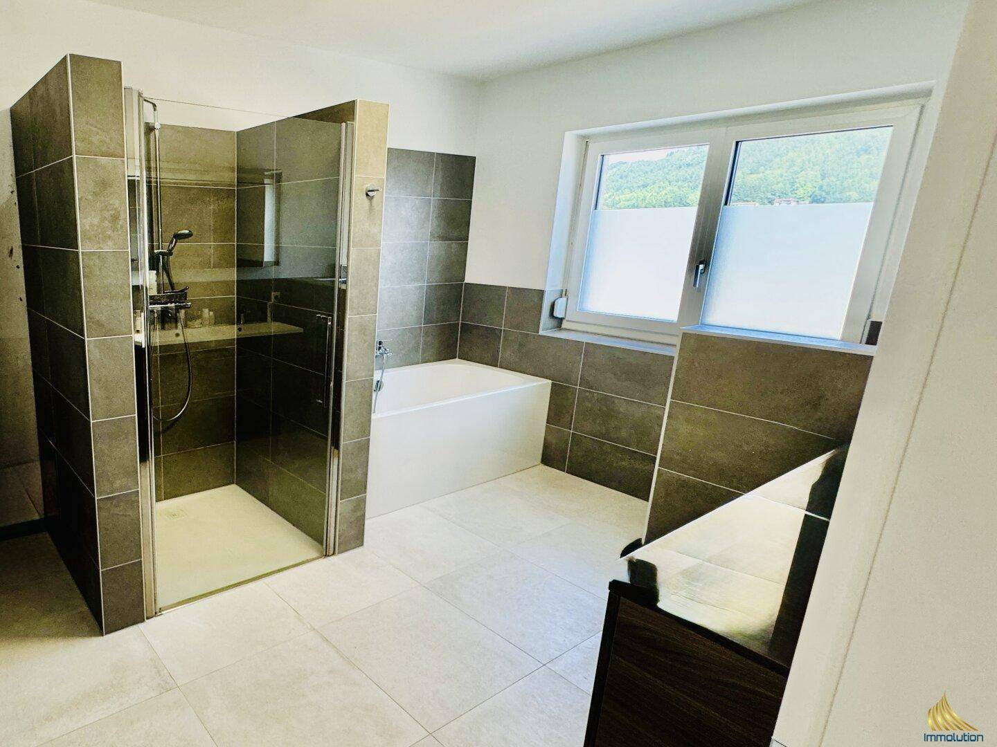 Modernes Badezimmer mit grauen Fliesen, begehbarer Dusche, Badewanne und Fenster.