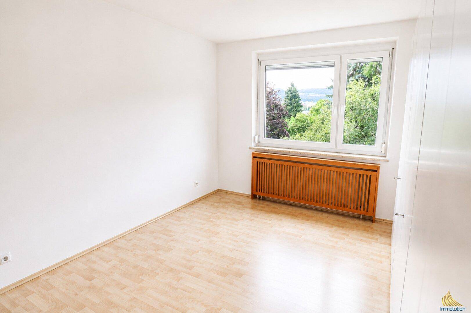 Helles Schlafzimmer mit Holzboden, Fenster und einem Heizkörper mit Holzverkleidung.