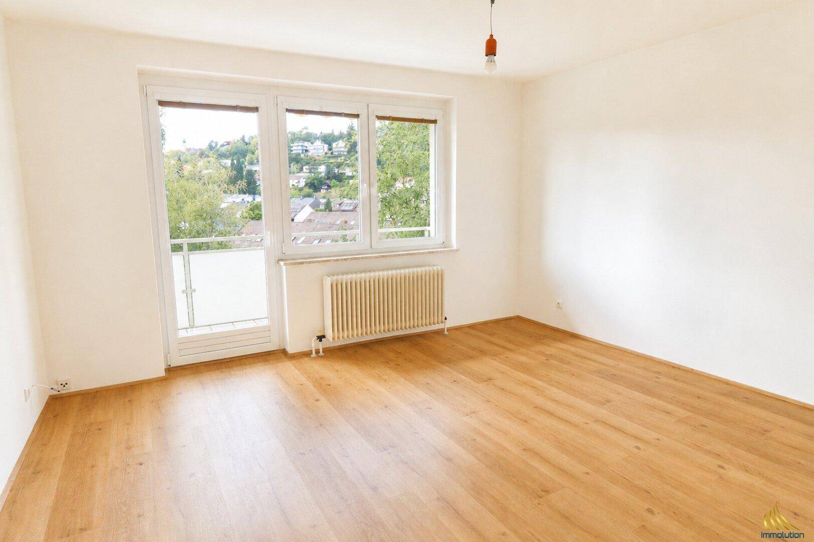 Helles Zimmer mit Holzboden, großem Fenster und Zugang zu einem kleinen Balkon.