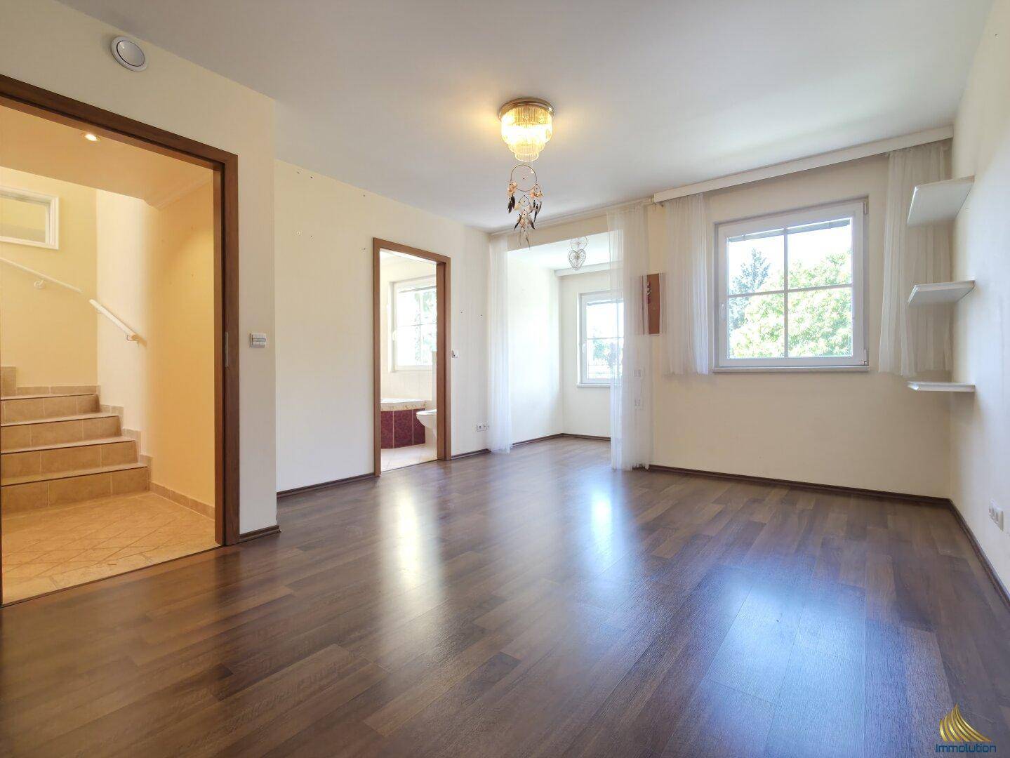 Heller Raum mit dunklem Holzboden, Fenstern und Zugang zum Treppenhaus.