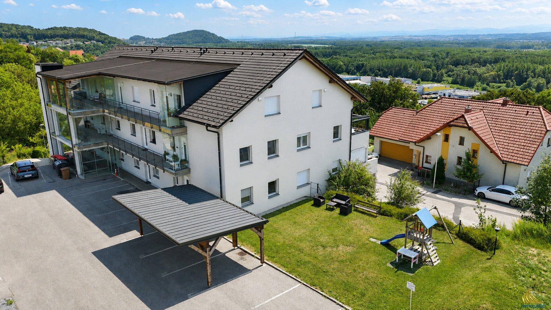 Mehrfamilienhaus mit Carport, Spielplatz und Grünflächen in ländlicher Umgebung.