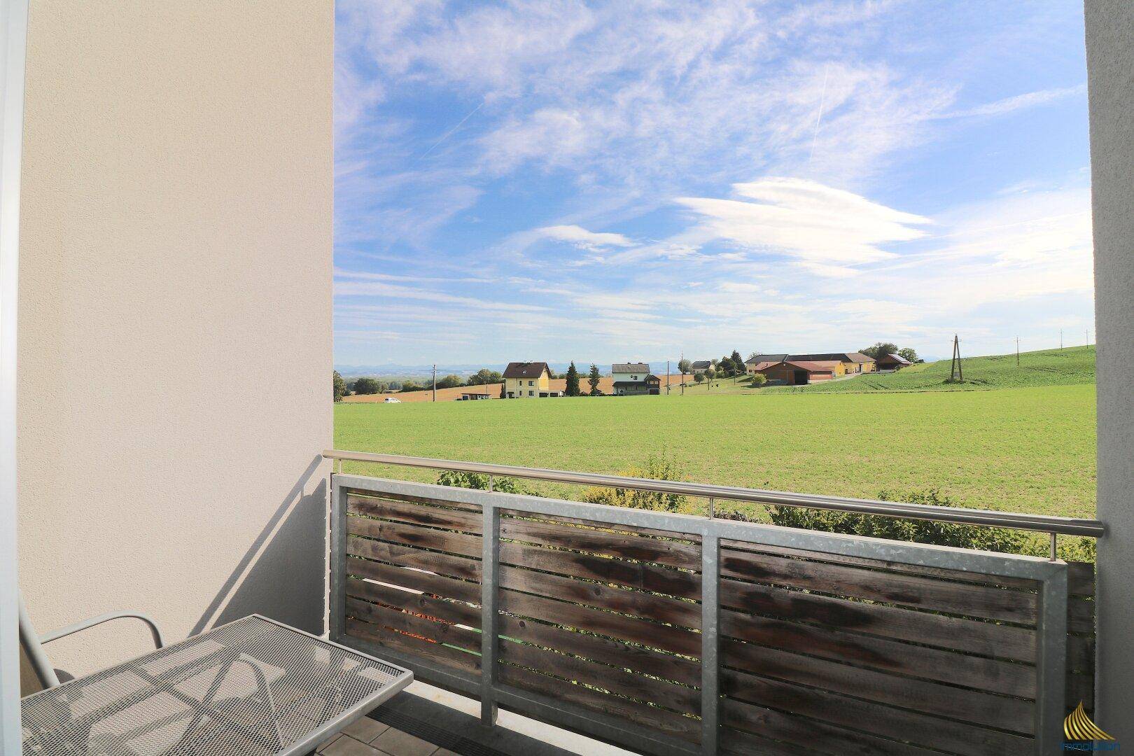 Balkon mit Tisch und Stühlen und weitem, sonnigem Blick über die grüne Landschaft.