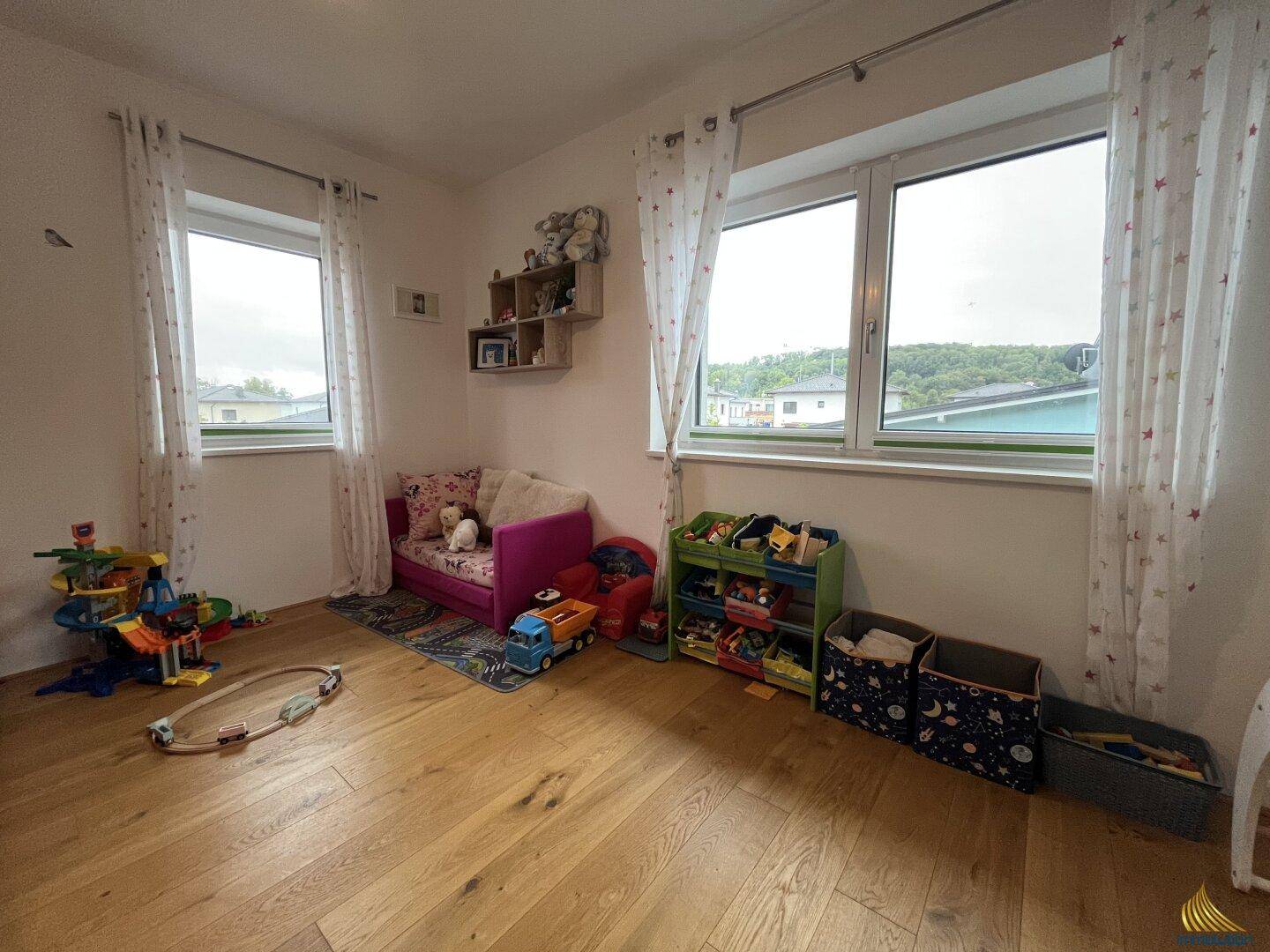 Helles Kinderzimmer mit Holzboden, zwei Fenstern und viel Platz zum Spielen.