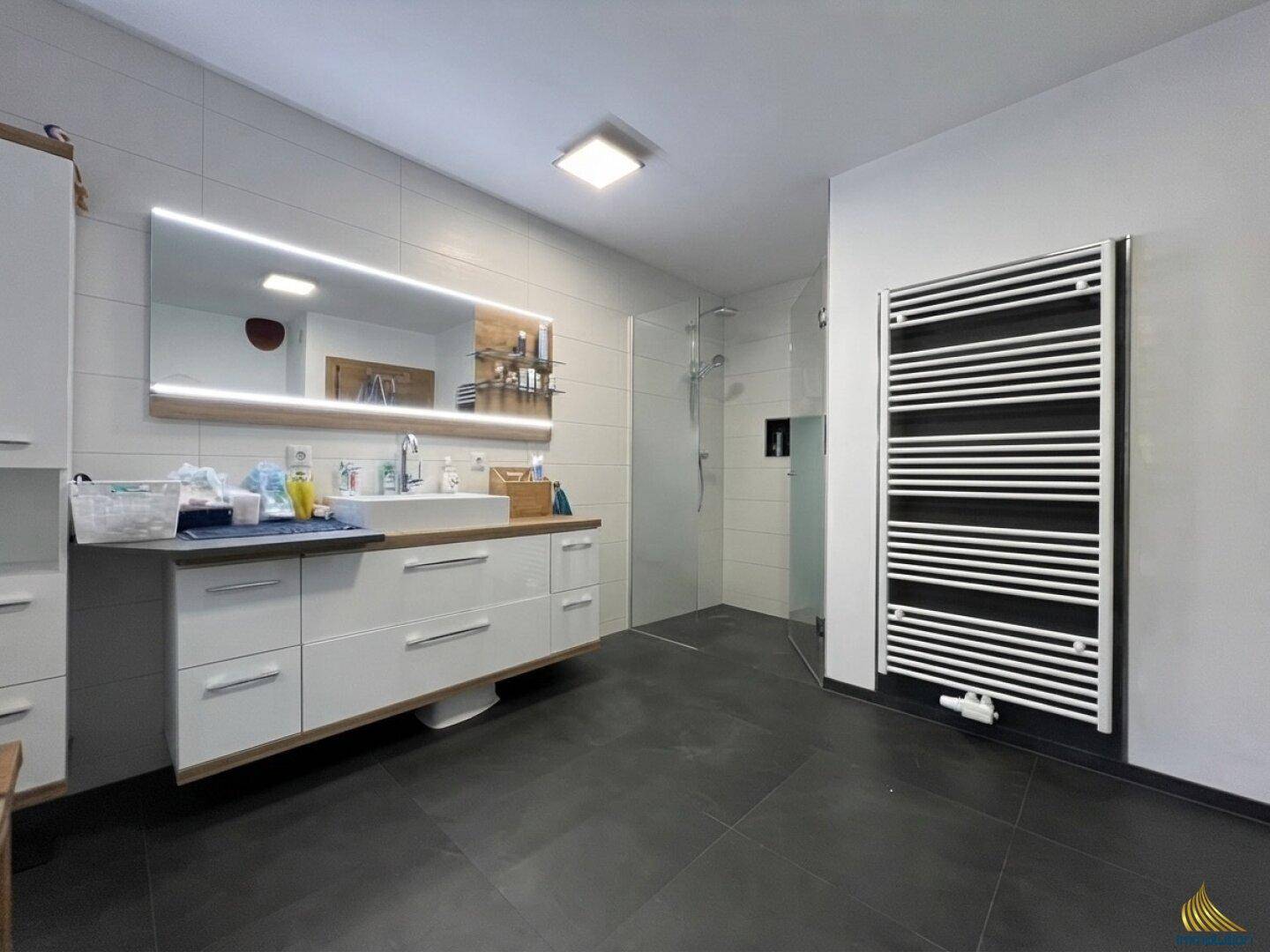 Elegantes Badezimmer mit großem Waschtisch, beleuchtetem Spiegel und begehbarer Dusche sowie Handtuchheizkörper.