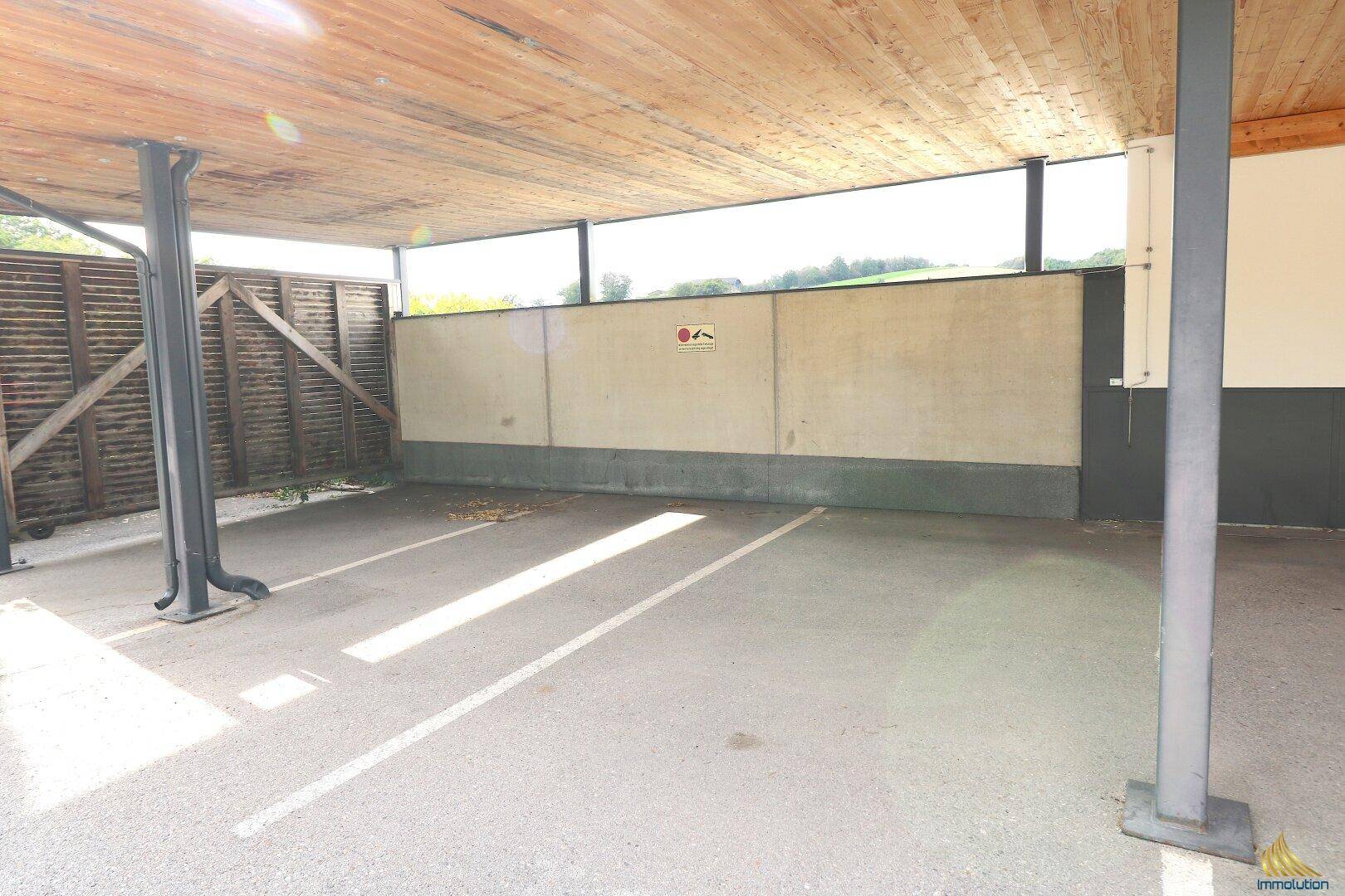 Überdachter Parkplatz mit Betonboden und Holzdecke, bietet Schutz für Fahrzeuge.