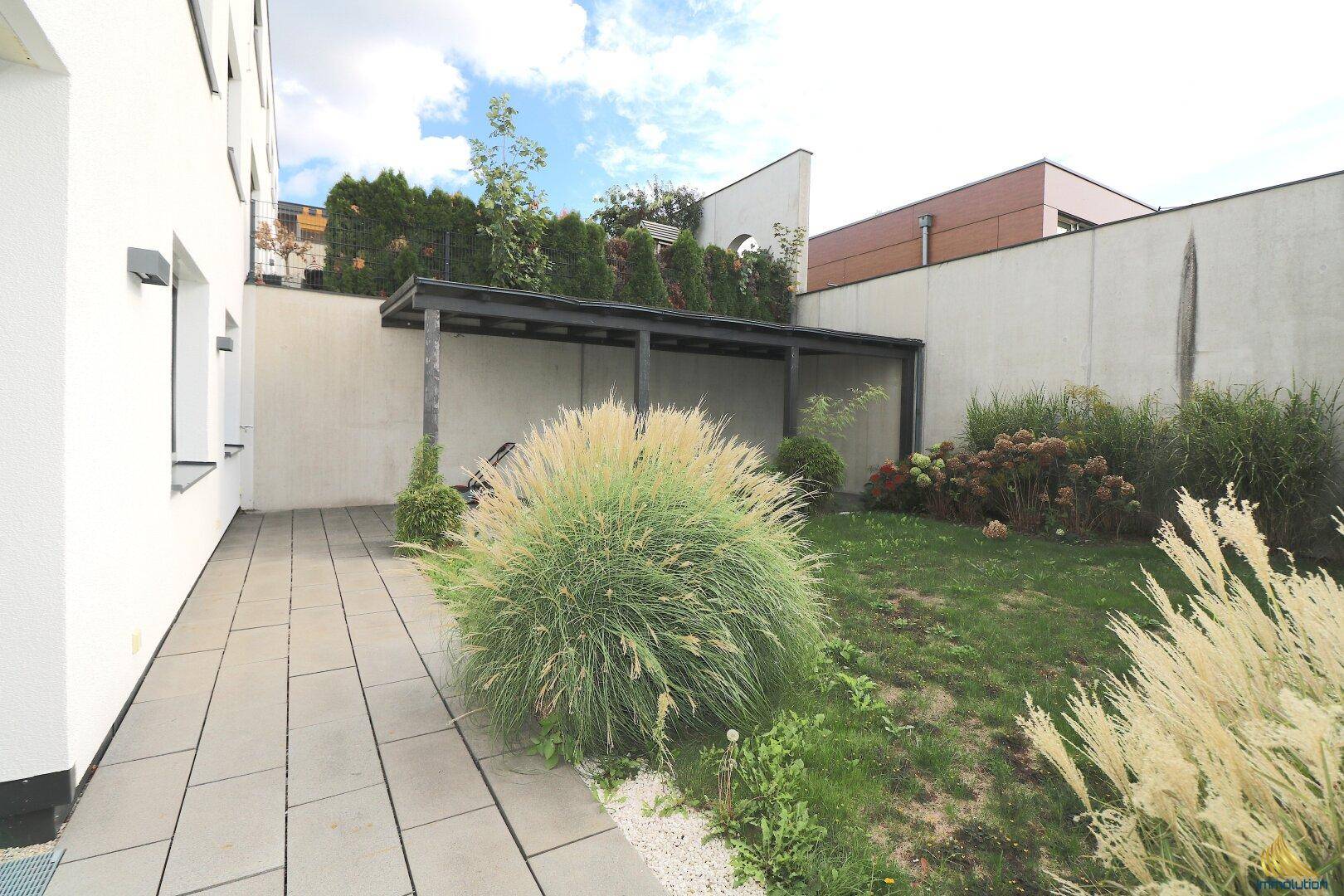 Gepflasterte Terrasse mit Gartenbereich, üppiger Bepflanzung und einer überdachten Sitzgelegenheit.