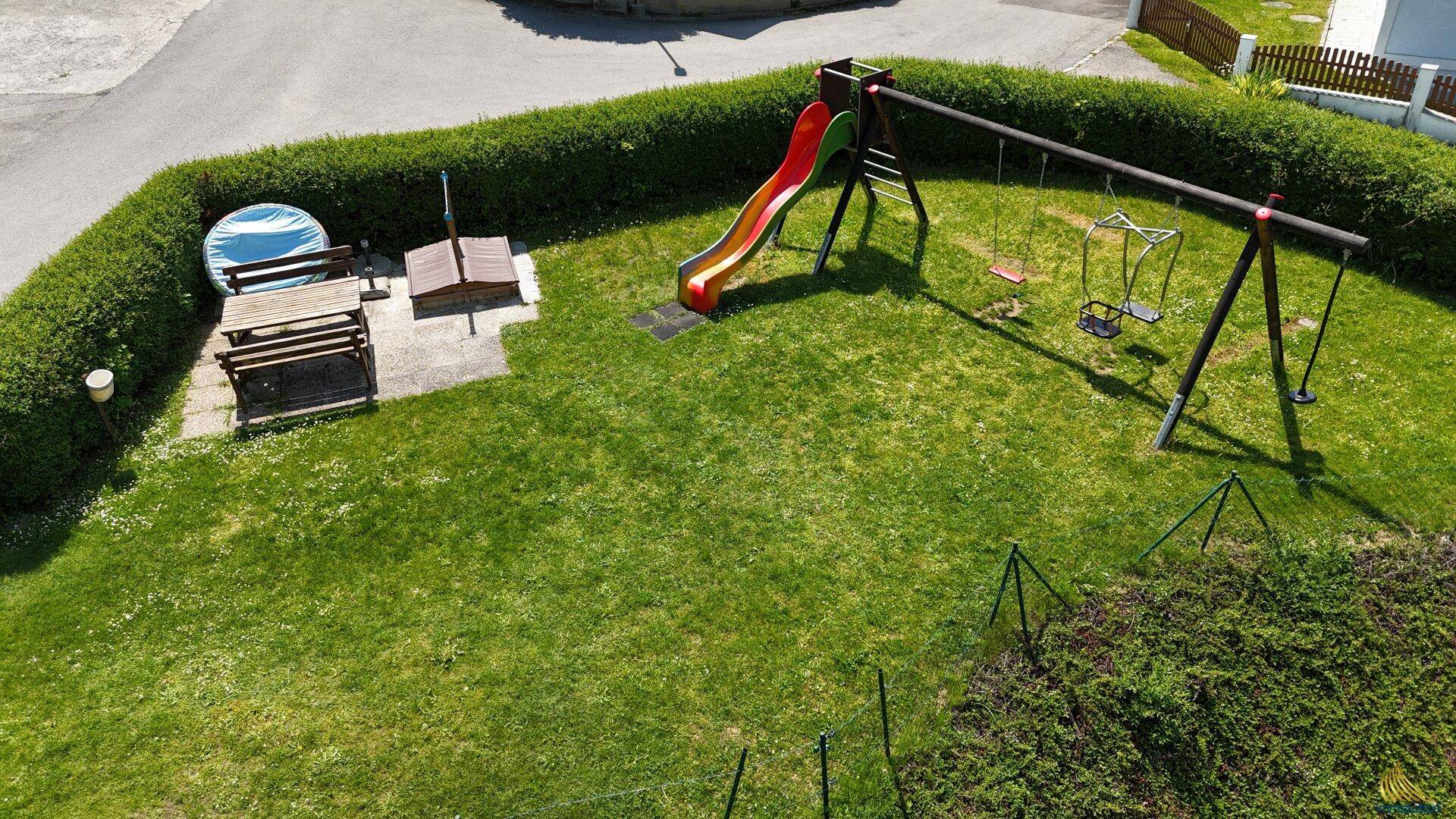Spielplatz mit Rutsche, Schaukeln, Sandkasten und Picknicktisch, umgeben von einer Hecke.