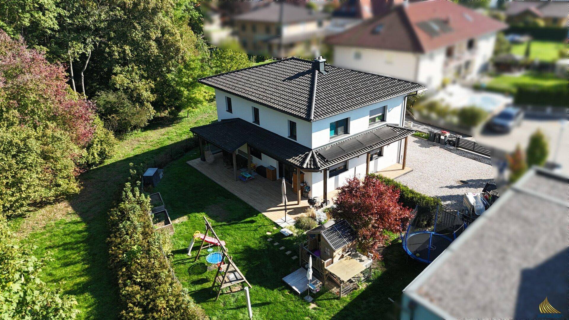 Rückansicht des Hauses mit überdachter Holzterrasse, weitläufigem Rasen und Kinderspielplatz im Grünen.
