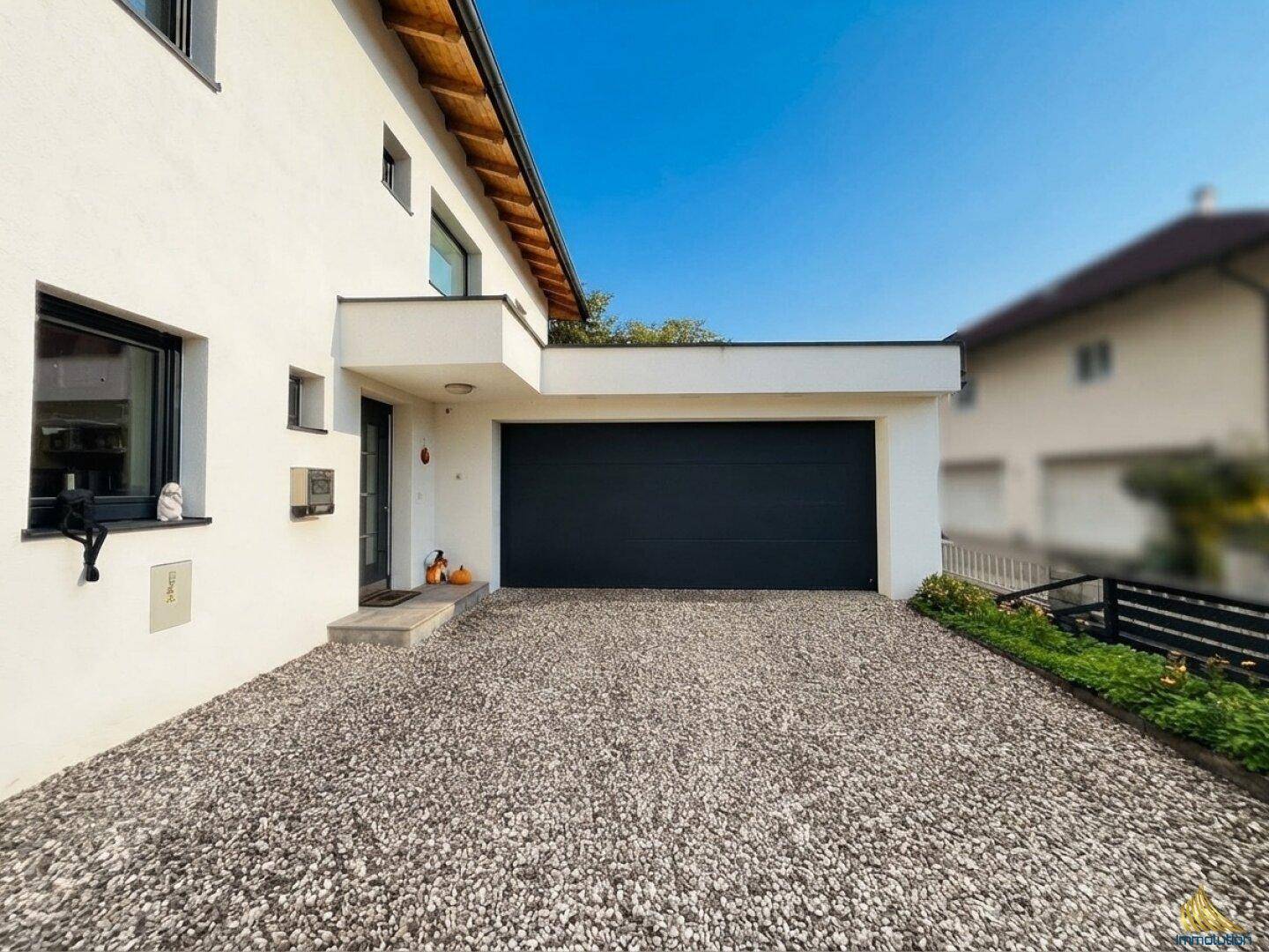 Vorderansicht des Hauses mit modernem Eingangsbereich, dunklem Garagentor und Kiesauffahrt.