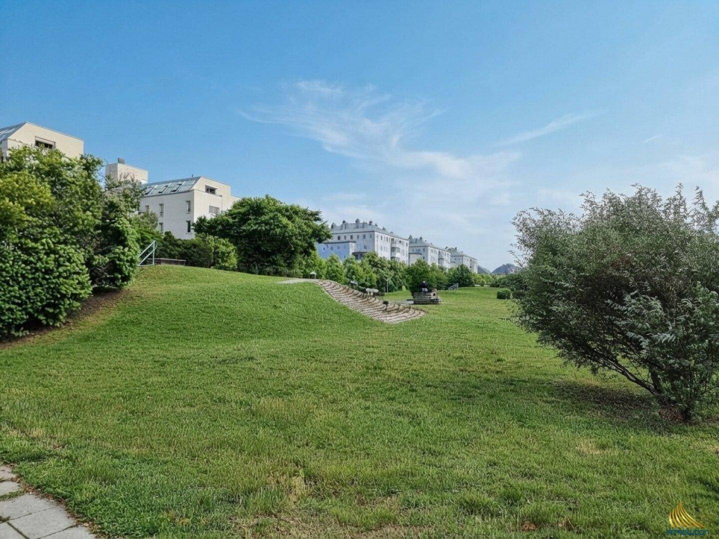 Weitläufige Grünfläche mit sanfter Neigung, Sitzbänken und Wohngebäuden in der Ferne unter blauem Himmel.