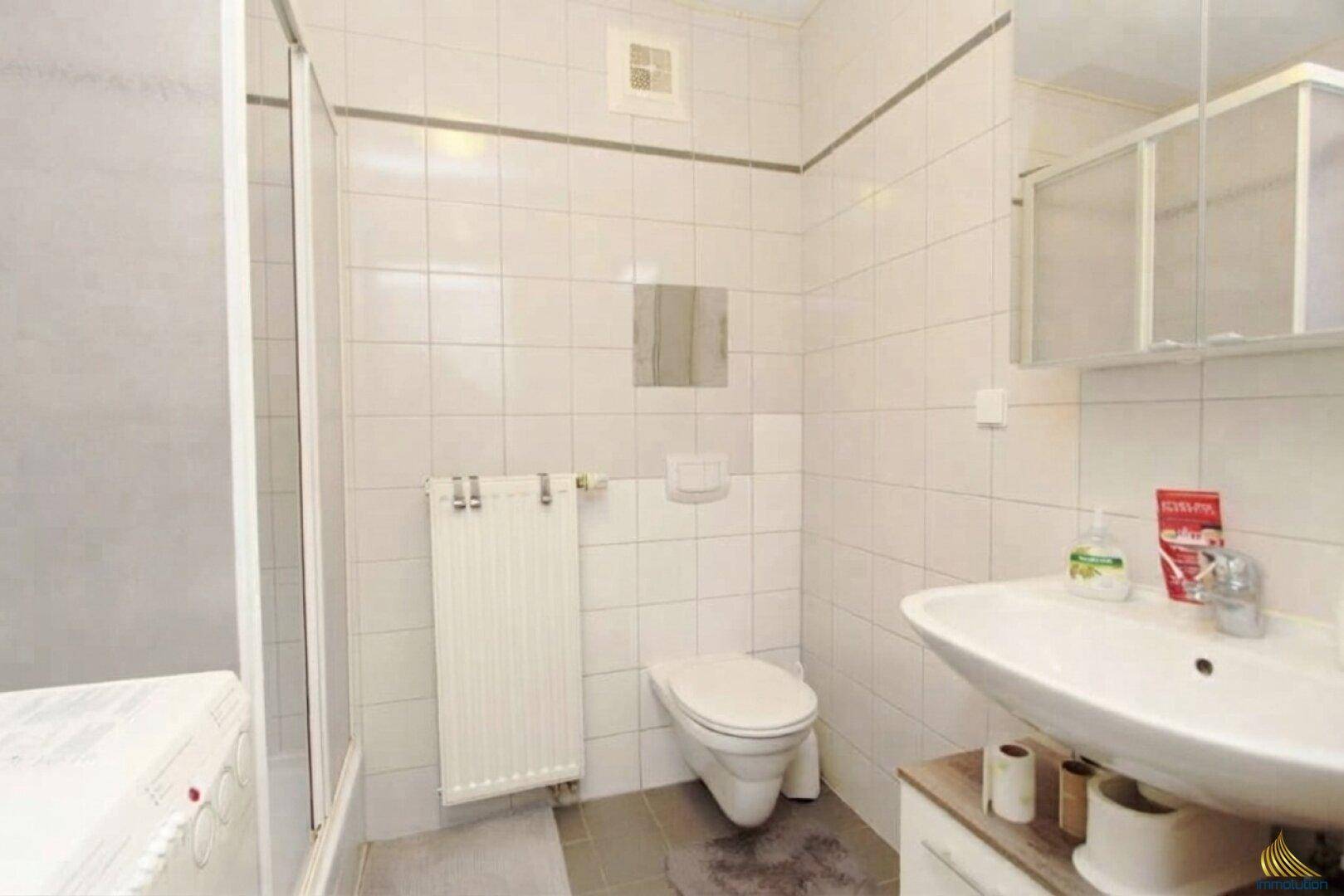 Badezimmer mit weißen Fliesen, Toilette, Waschbecken, Duschbereich und beheiztem Handtuchhalter.