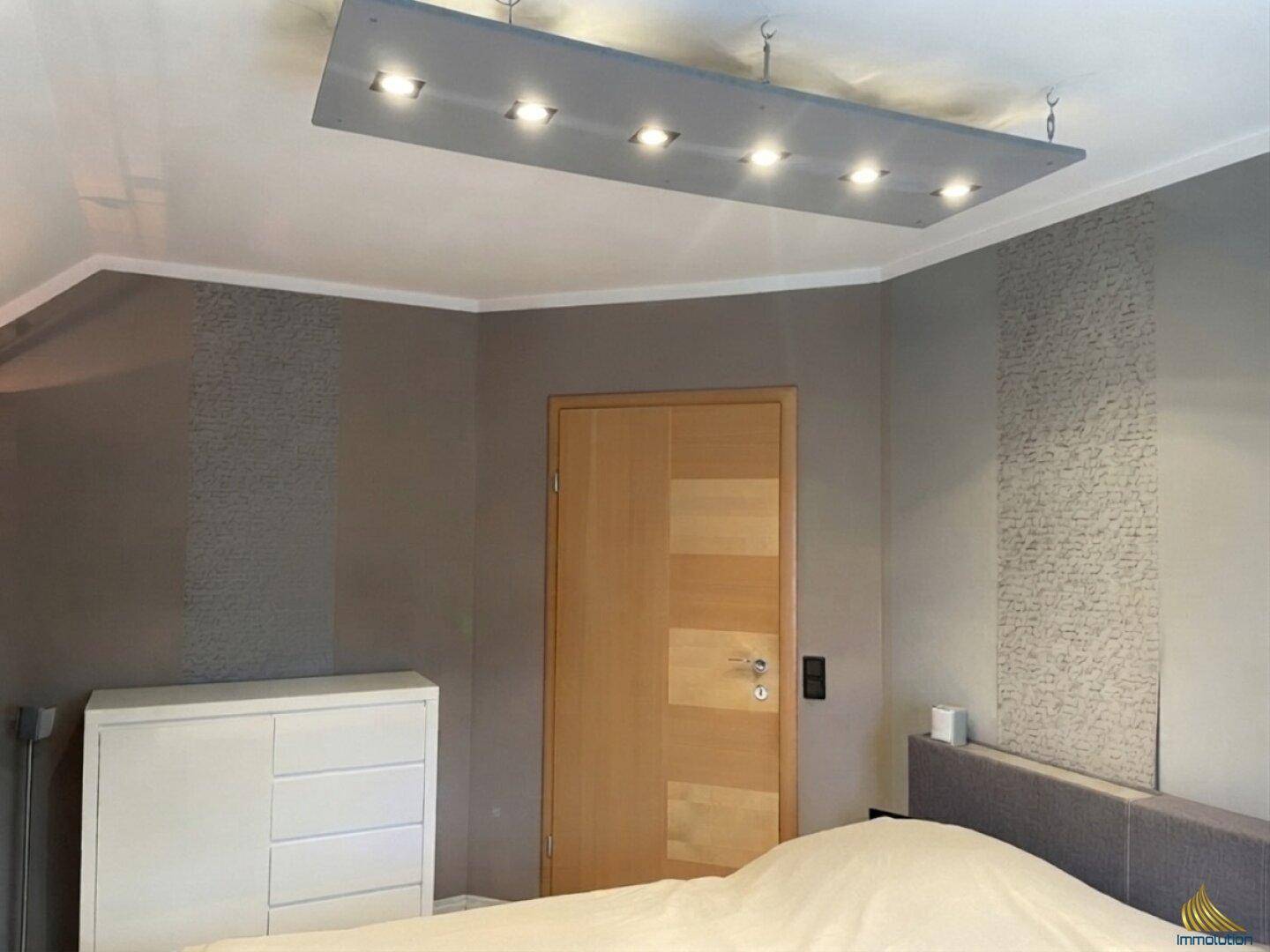 Schlafzimmer mit Holztür, weißer Kommode und indirekter Deckenbeleuchtung.