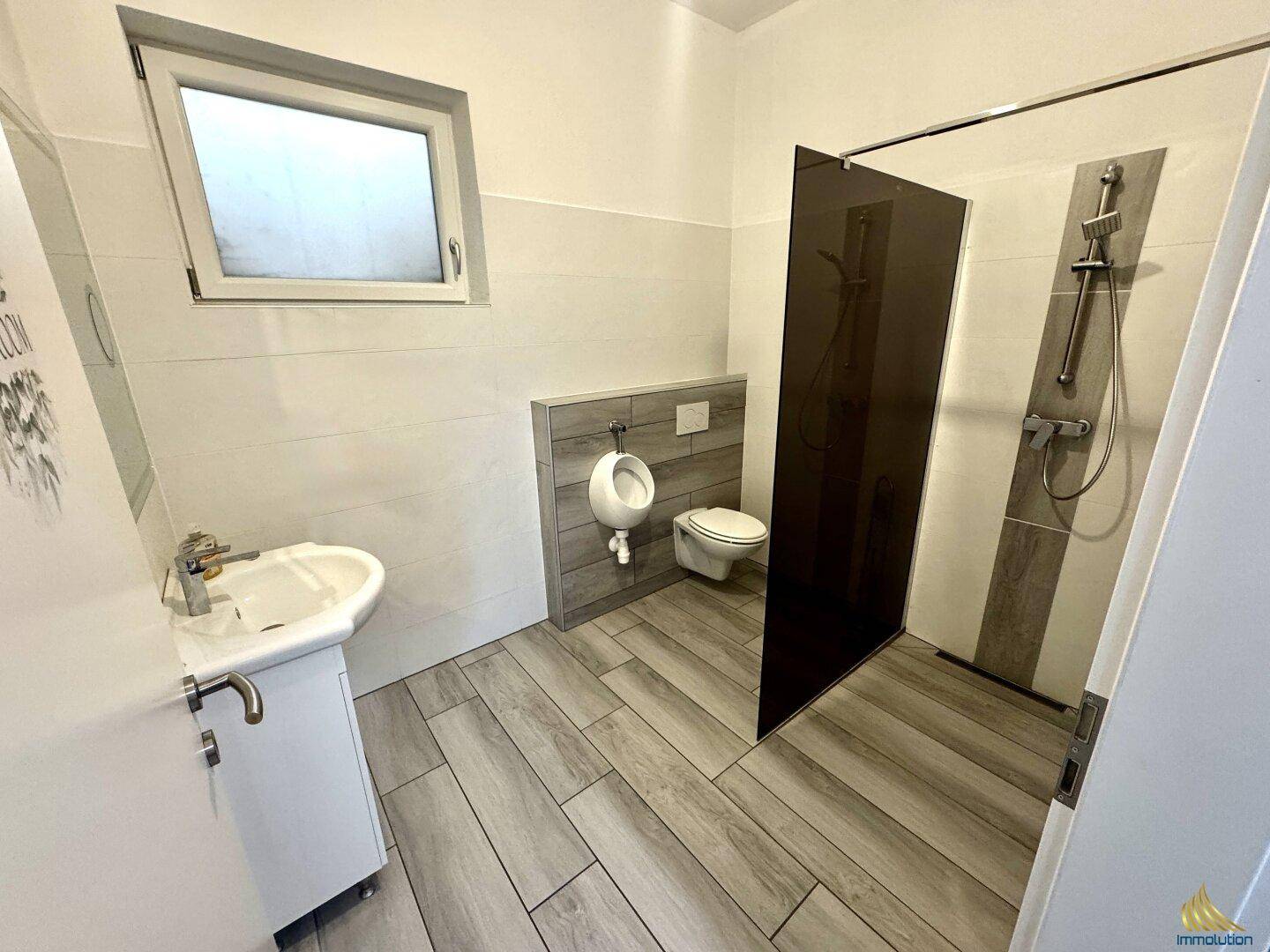 Badezimmer mit Dusche, WC, Urinal und Fenster, modern gefliest und hell.