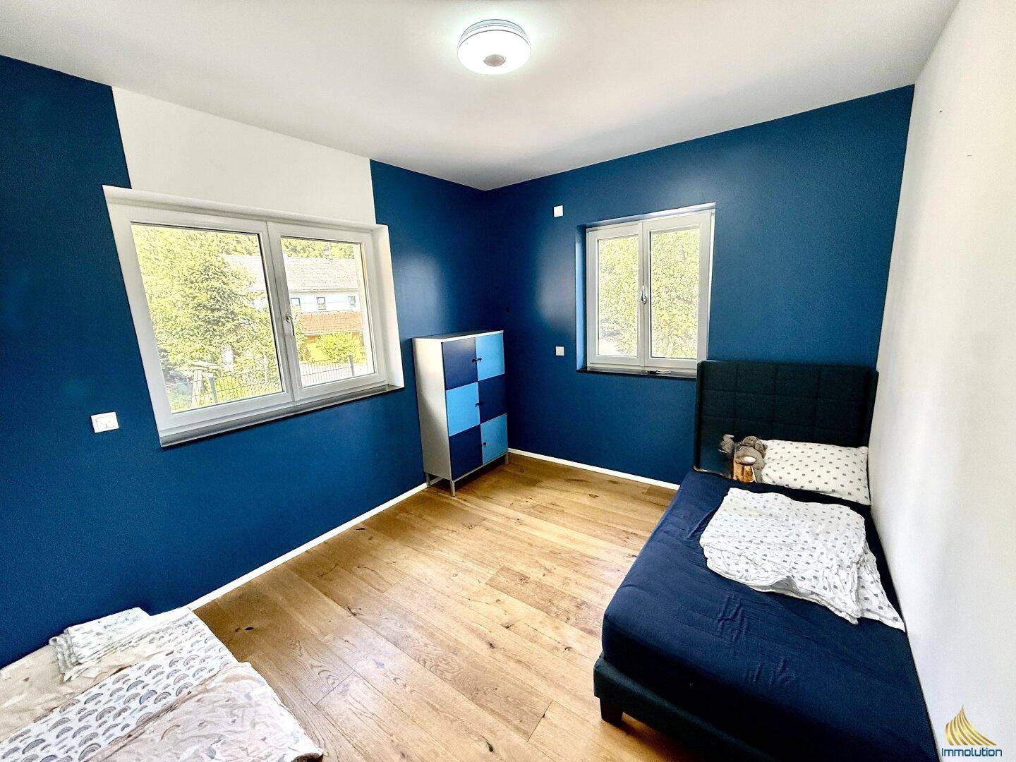 Kinderzimmer mit blauen Wänden, Holzboden und zwei Fenstern für viel Tageslicht.