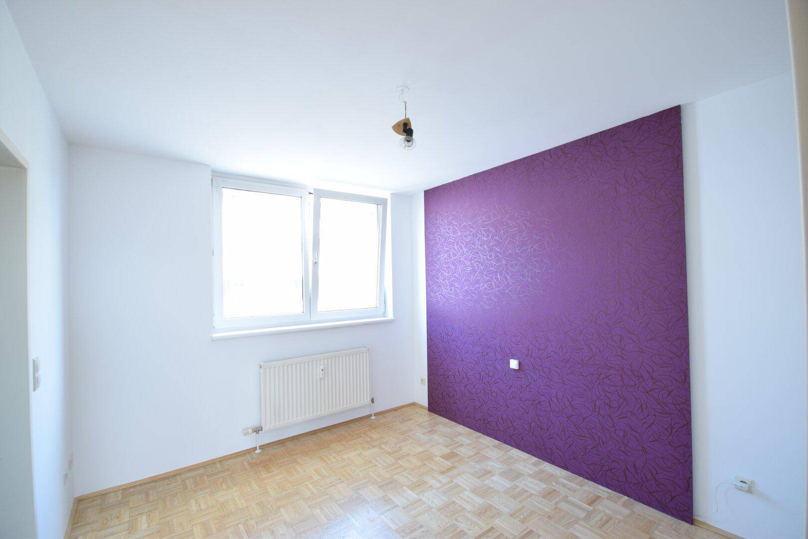 Schlafzimmer mit Parkettboden, einer auffälligen lila Akzentwand und einem Fenster für Tageslicht.