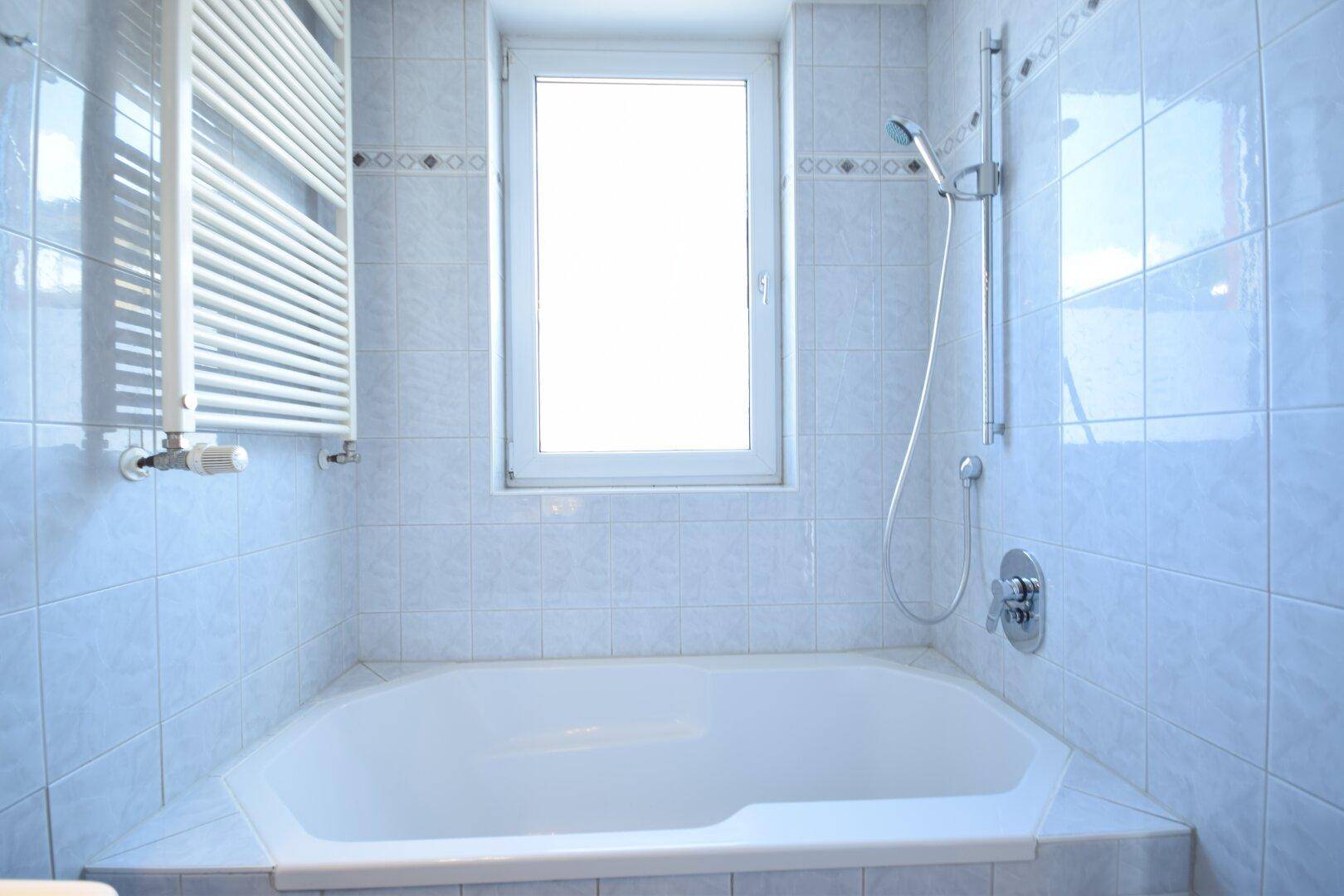 Badezimmer mit hellblauen Fliesen, einer Eckbadewanne, Duscharmatur und einem beheizten Handtuchhalter.