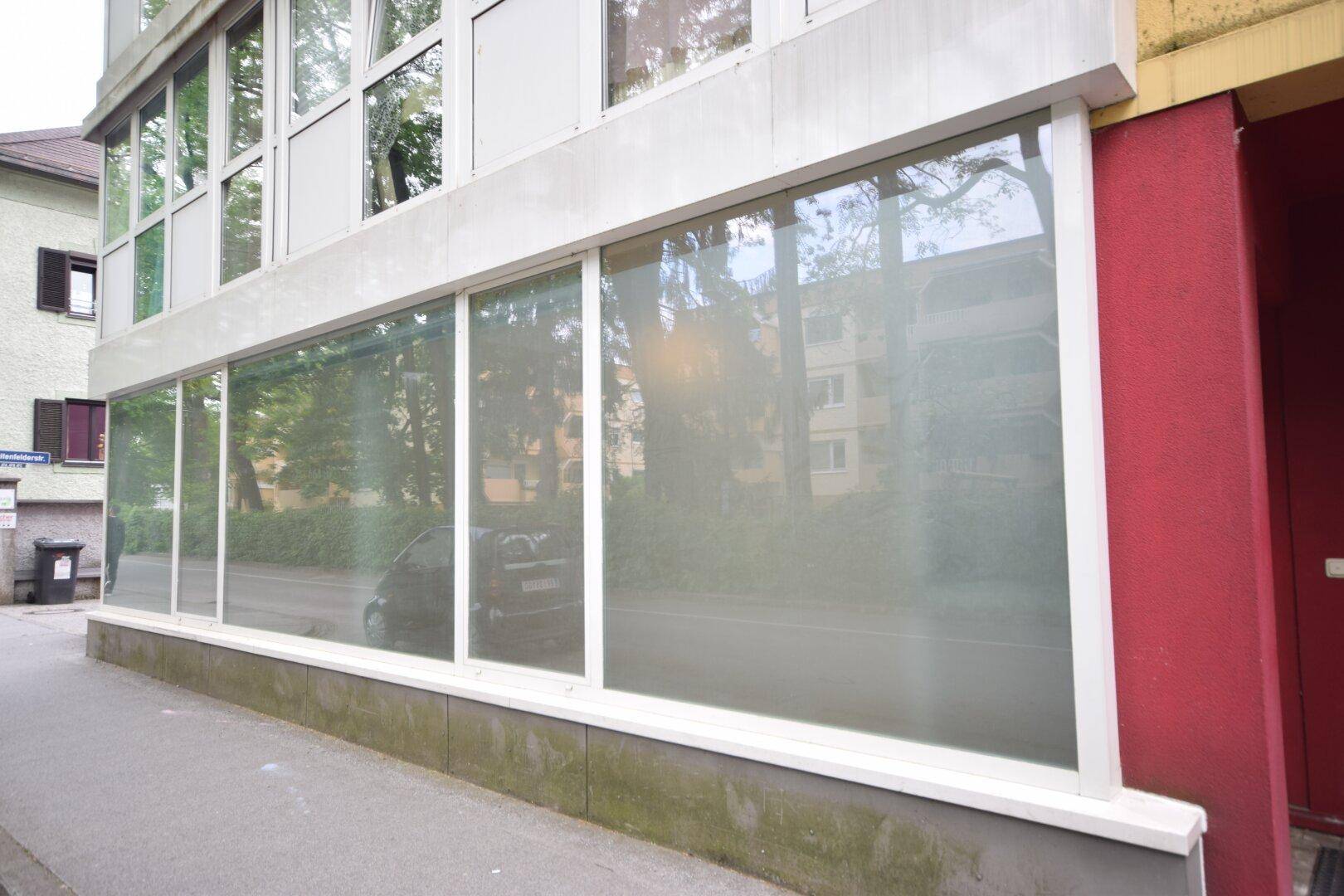 Gebäudefassade mit großen, undurchsichtigen Fenstern im Erdgeschoss und einem roten Gebäudeteil daneben.
