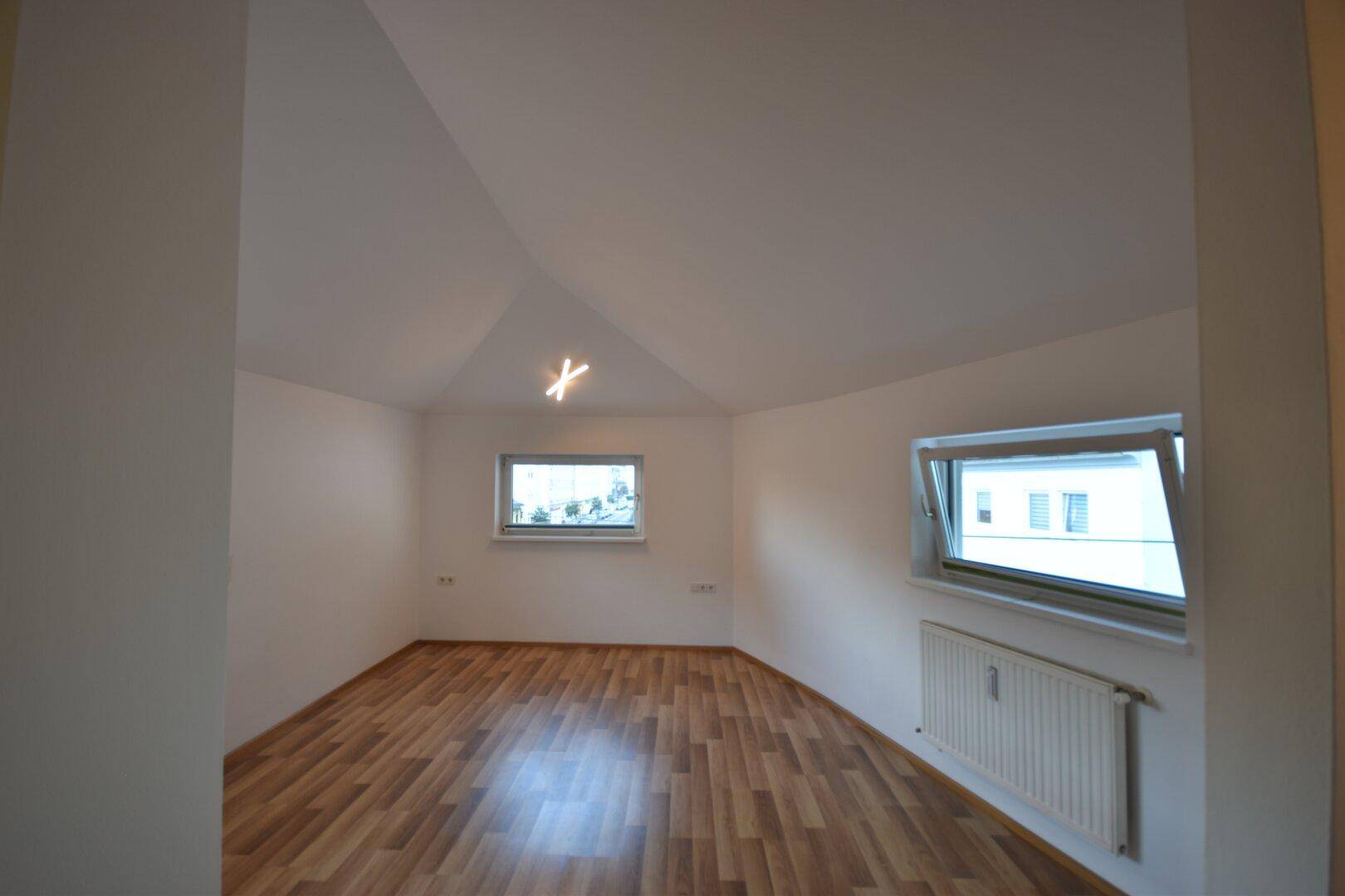 Heller Raum im Dachgeschoss mit Laminatboden, schrägen Decken und zwei Fenstern für natürliches Licht.