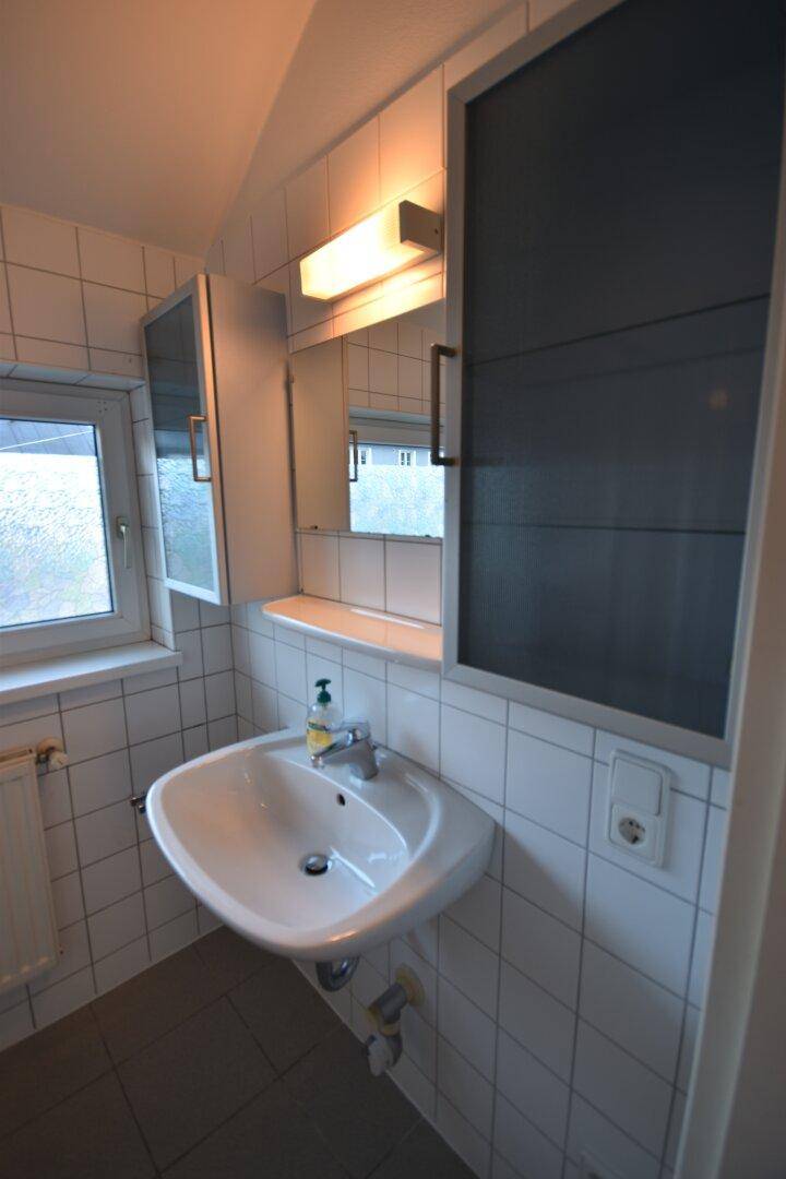 Funktionales Badezimmer mit weißem Waschbecken, Spiegelschrank, weißen Wandfliesen und Fenster.