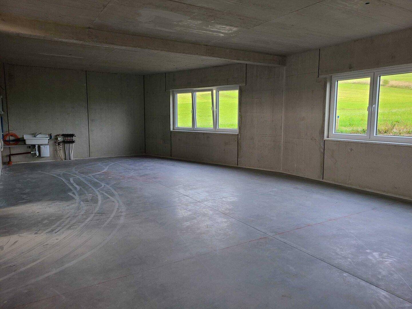 Heller Industrieraum mit Betonboden und -wänden, großen Fenstern und Blick ins Grüne sowie sichtbaren Installationen.