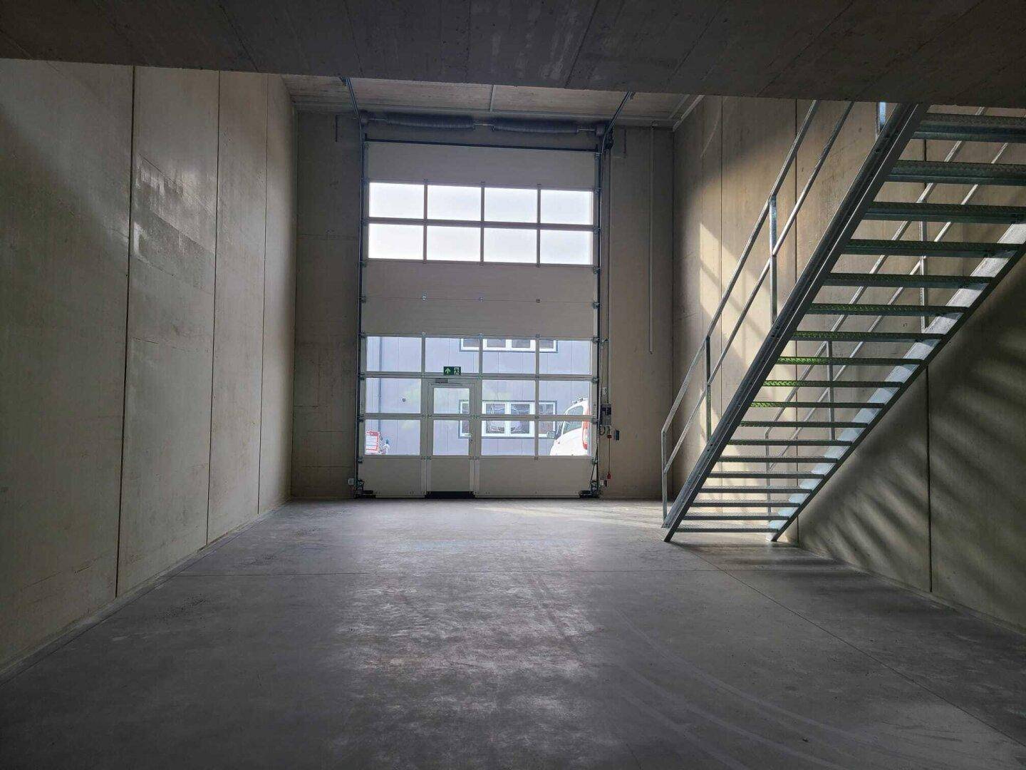 Großer Industrieraum mit Betonwänden und -boden, einer Metalltreppe und einem hohen Rolltor am Ende des Raumes.
